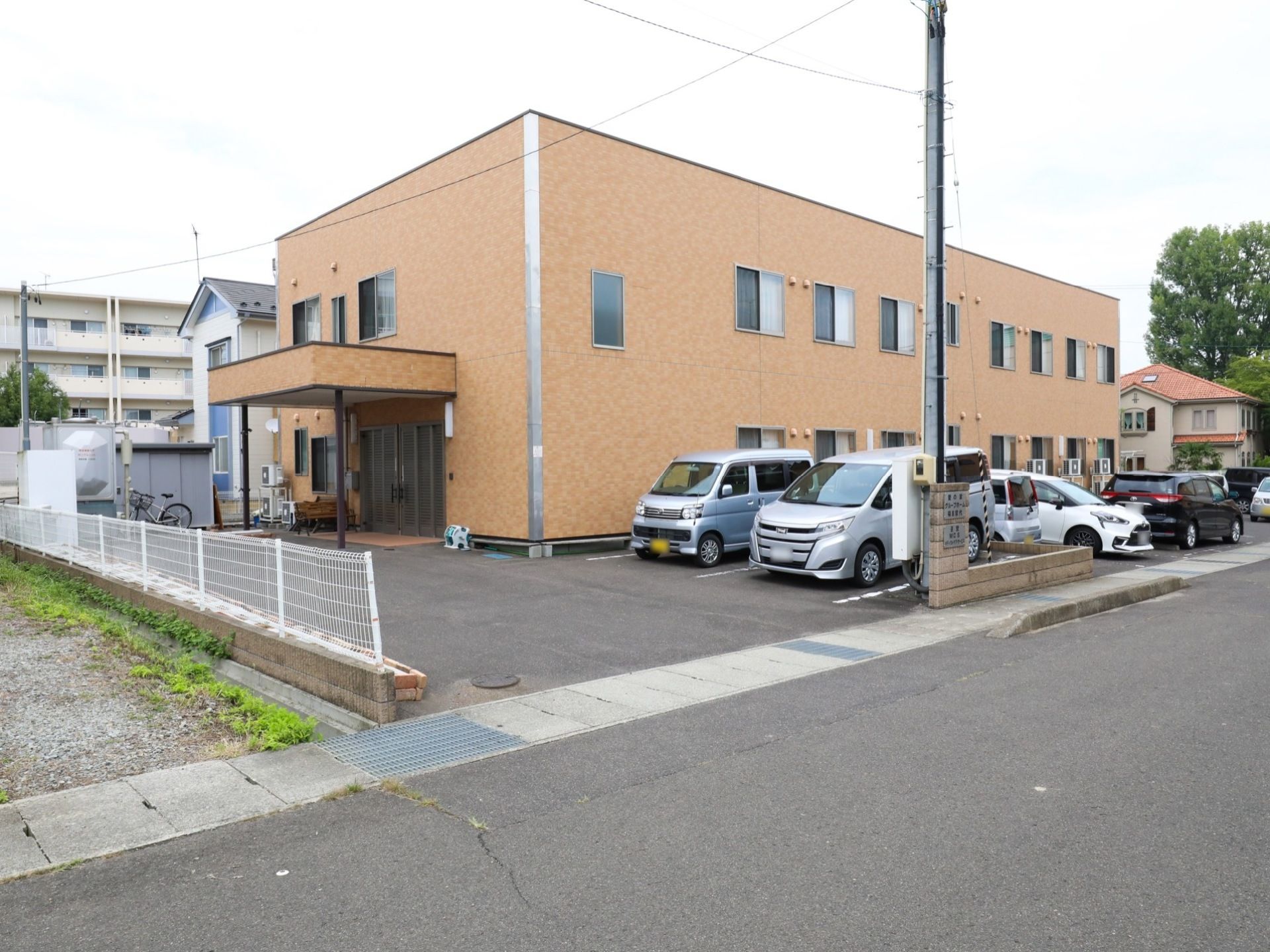 施設の外観は、２階建てでボックス型の建物である。玄関の前は、ゆったりとしたスペースがあり、門と建物の間に数台の車が駐車している。