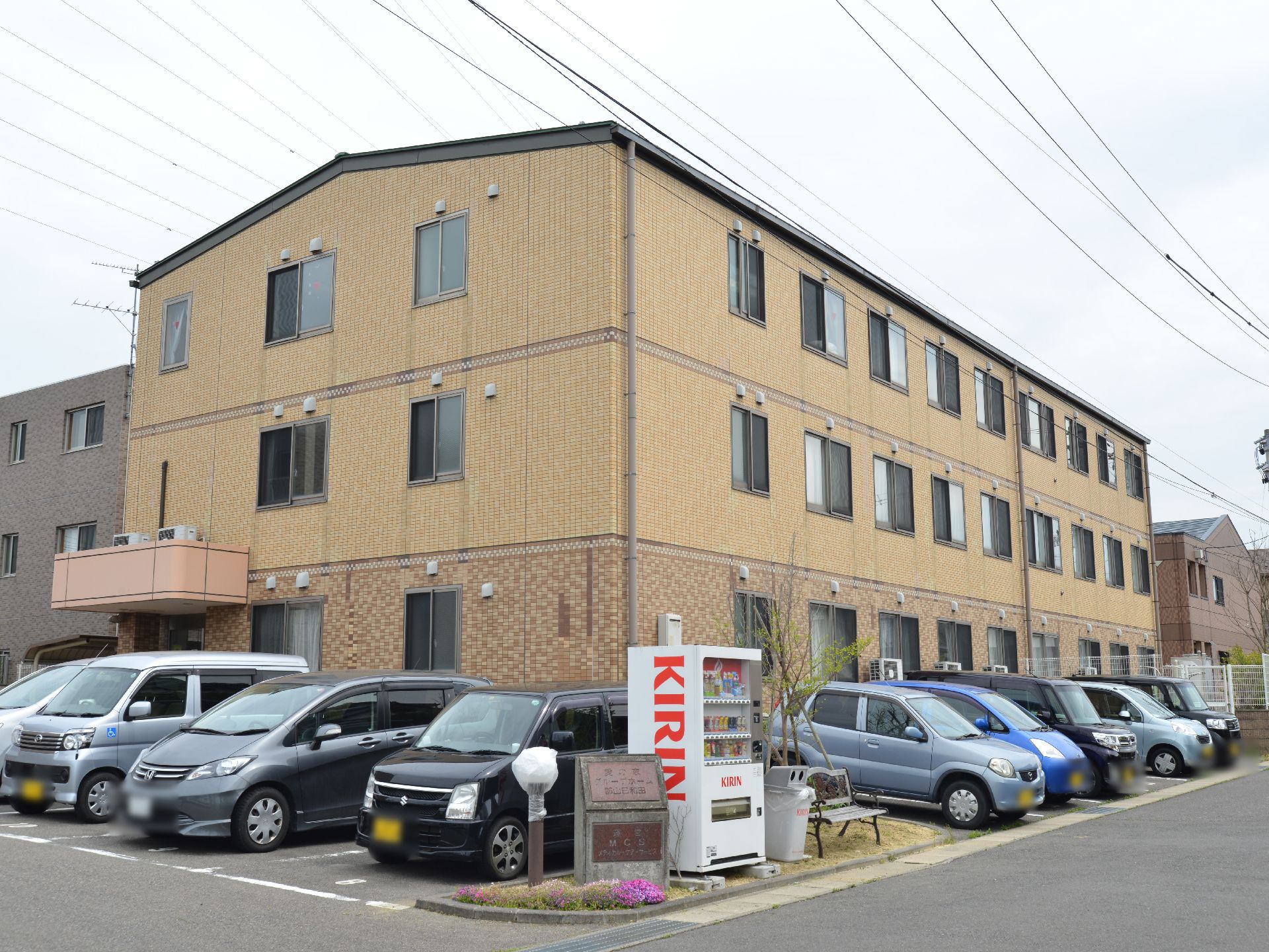 施設は3階建てである。色は濃いベージュ系。建物を取り囲むように駐車スペースがある。自動販売機のそばにはベンチがある。