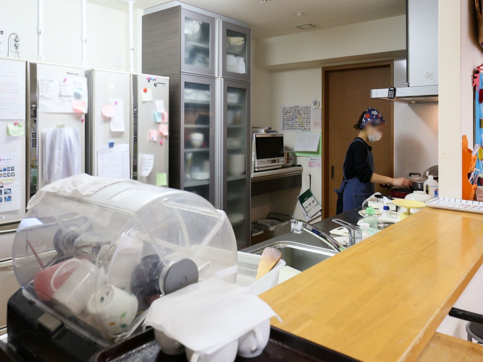 右手前に木製のカウンターがあるキッチンの図である。奥の壁に冷蔵庫や食器棚が並び、スタッフが作業をしている。