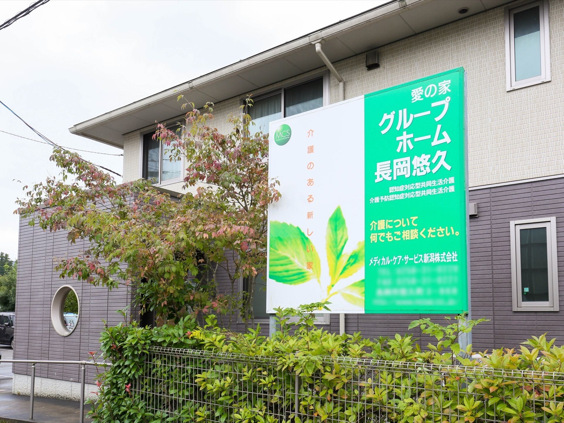 施設の外に立っている看板が中央にあり、その前に植え込みがある。奥に施設が見えており、１階がグレー、２階がベージュに塗られている。