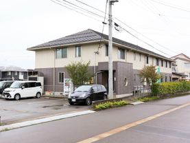 ２階建ての施設が中央に建っている。施設の前は道路が通っている。入り口前に白い車と黒い車が停められている。