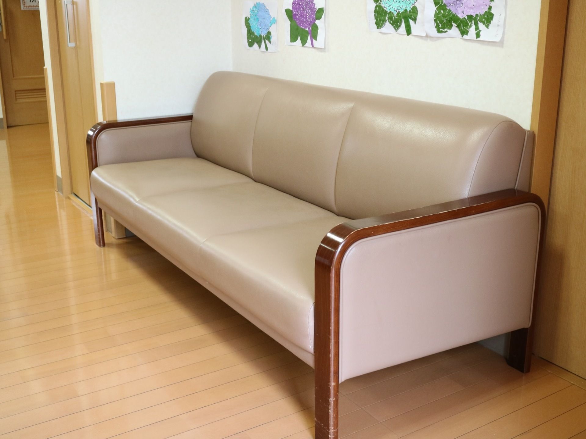 落ち着いた床色のフローリング部屋に、３人掛けのソファが置かれてある。壁には色とりどりの花が描かれた作品が貼られている。