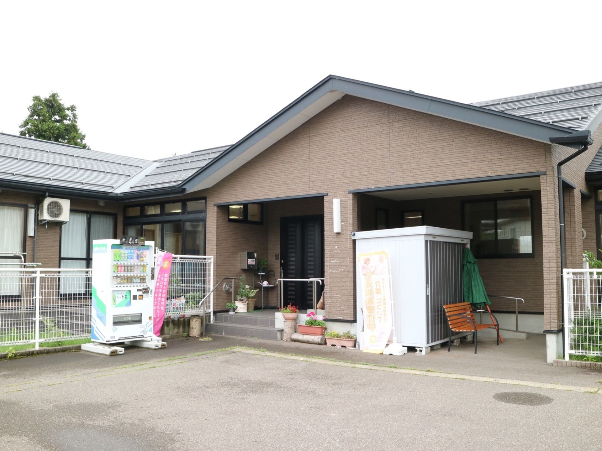 落ち着いた色合いの外観である。建物のまわりに、白色のフェンスが立っている。自動販売機も設置されてある。