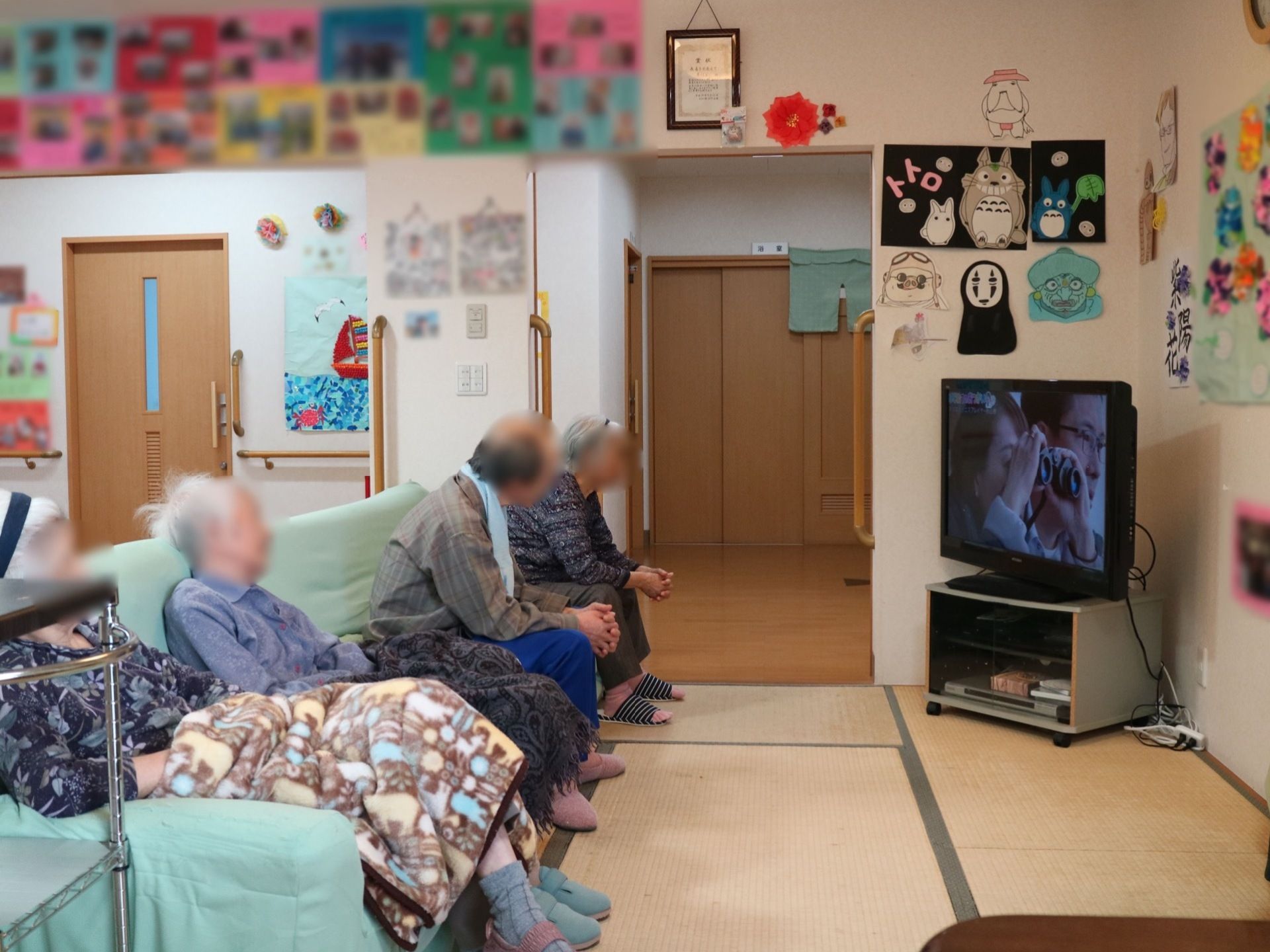 畳敷きのスペースに横長のソファーが置かれている。ソファーには緑色のカバーが掛けてある。部屋のすみにテレビがある。