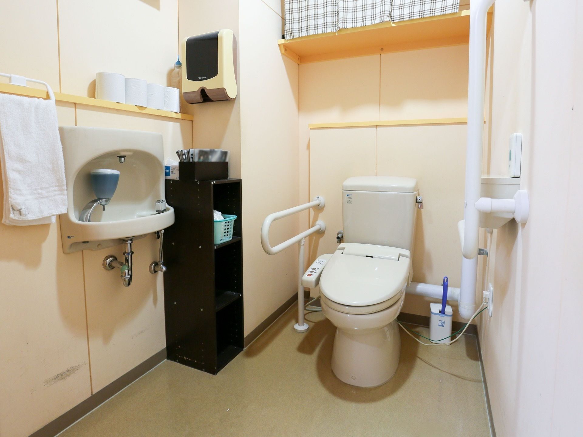 便器の周りには手すりや握り棒が配置されている。壁面には手洗いがあり、その上には予備のトイレットペーパーが準備されている。