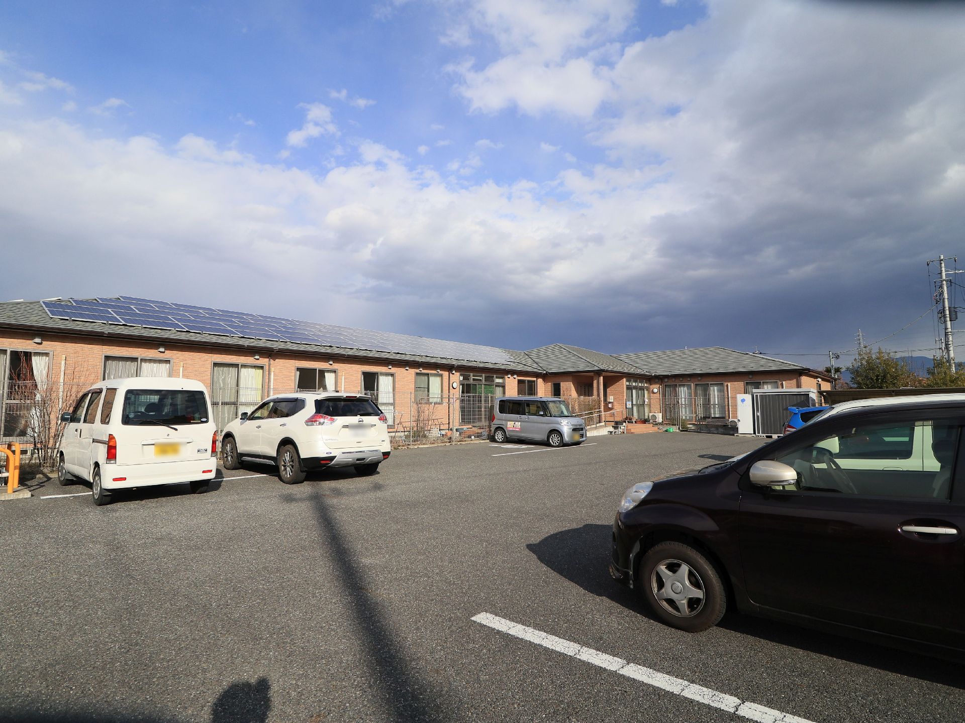 天気の良い日に、駐車場から施設を見た様子である。施設が完備している駐車場には、6台の車が停車している。