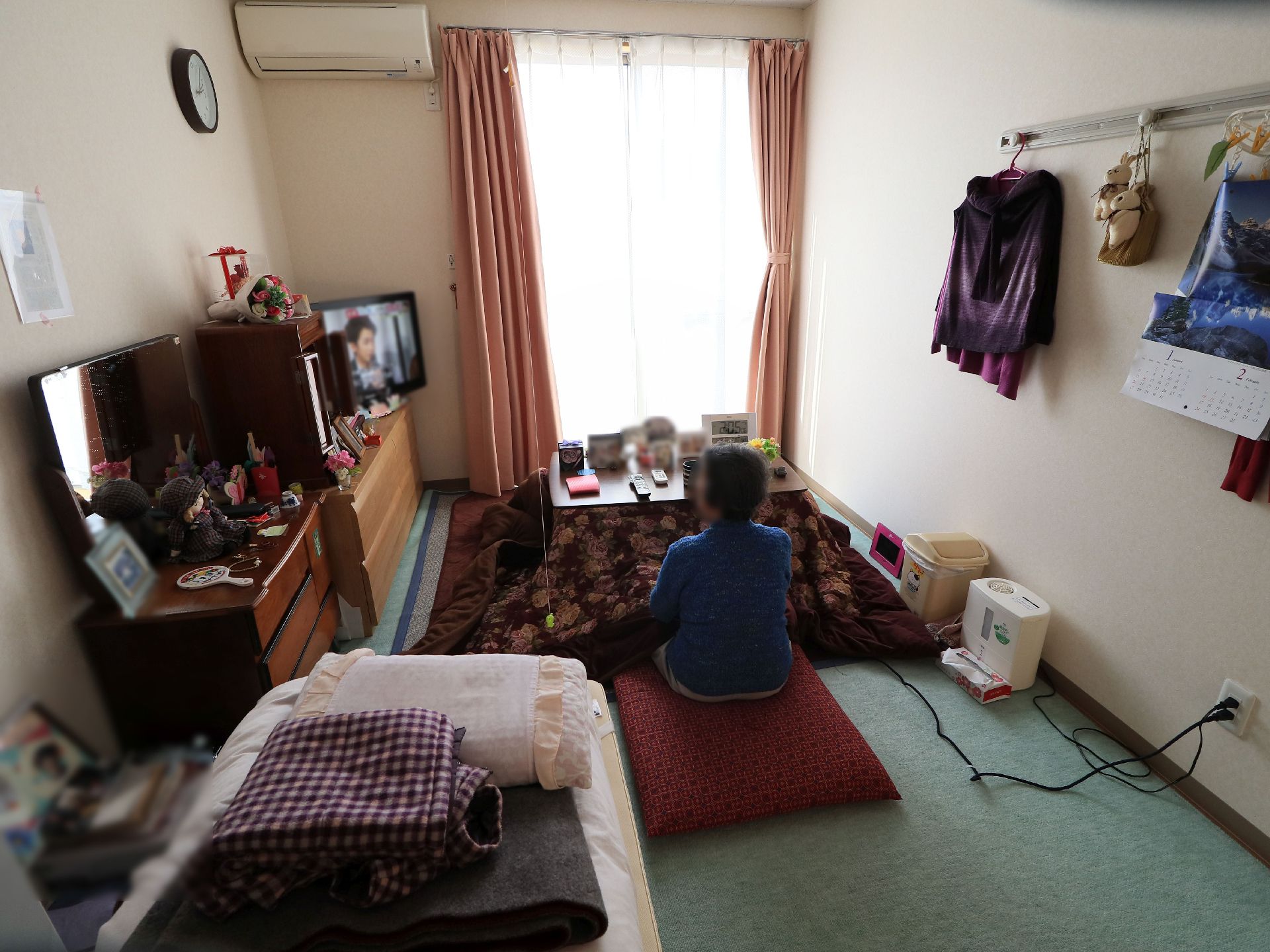 高さのある窓やエアコンが完備されている居室には、こたつに入りテレビをご覧になっている女性の入居者様の姿が見える。