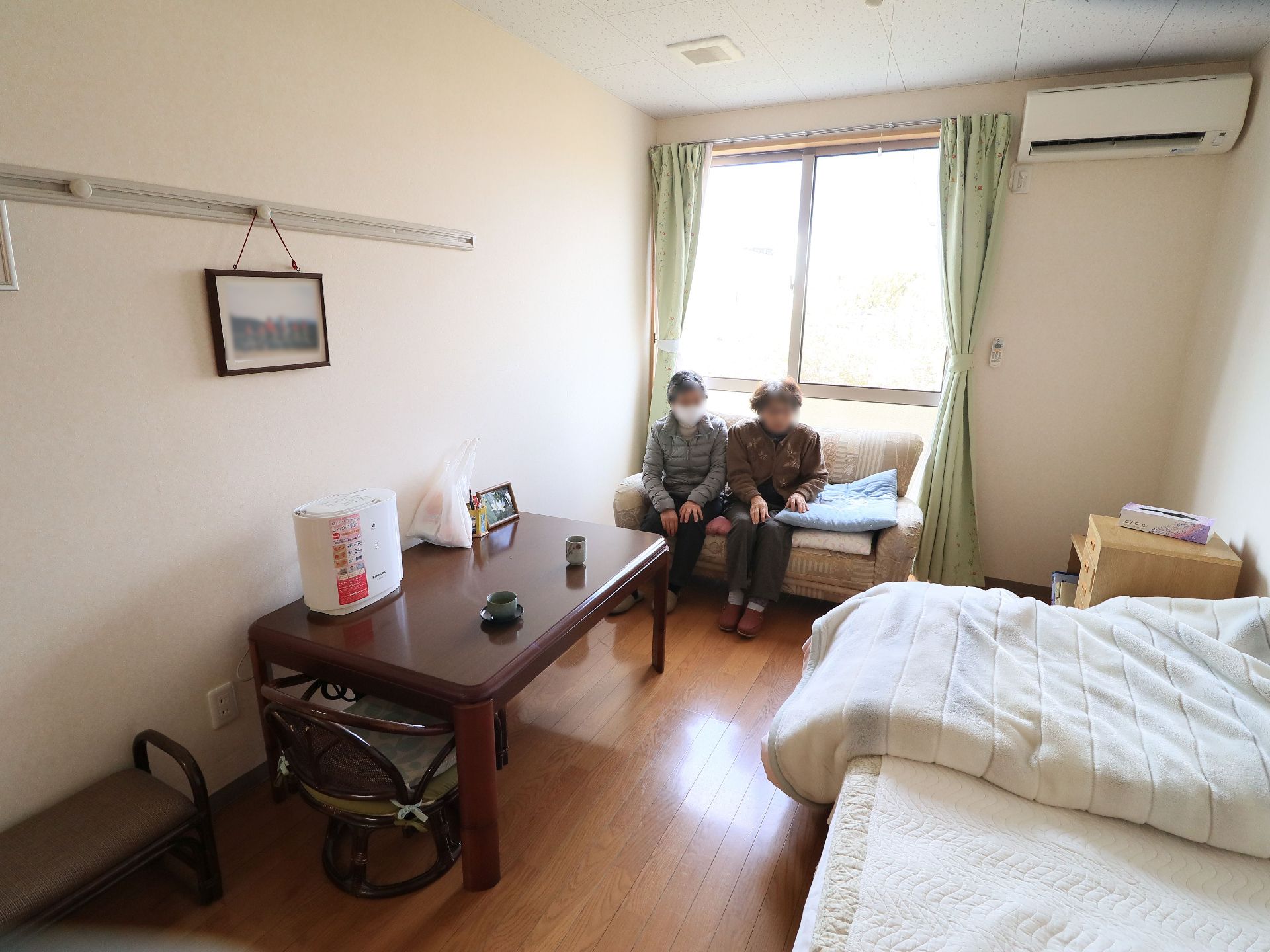 居室にはグリーンのカーテンのかかった窓がある。その前にソファが置かれていて、二人の入居者様が並んで座っている。
