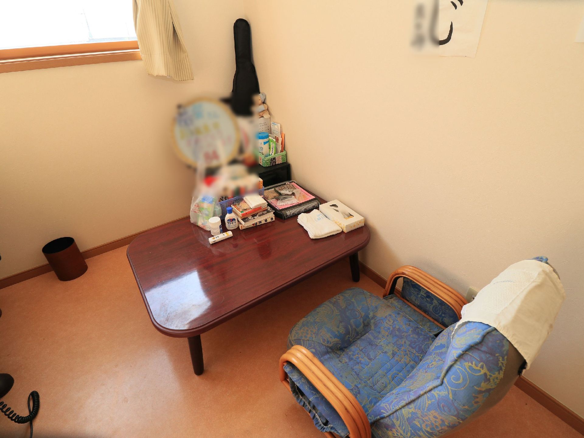 居室の一角に木目調のテーブルとブルーの座椅子が設置されている。テーブルの上にはティッシュやタオル、買い物袋などが置かれている。