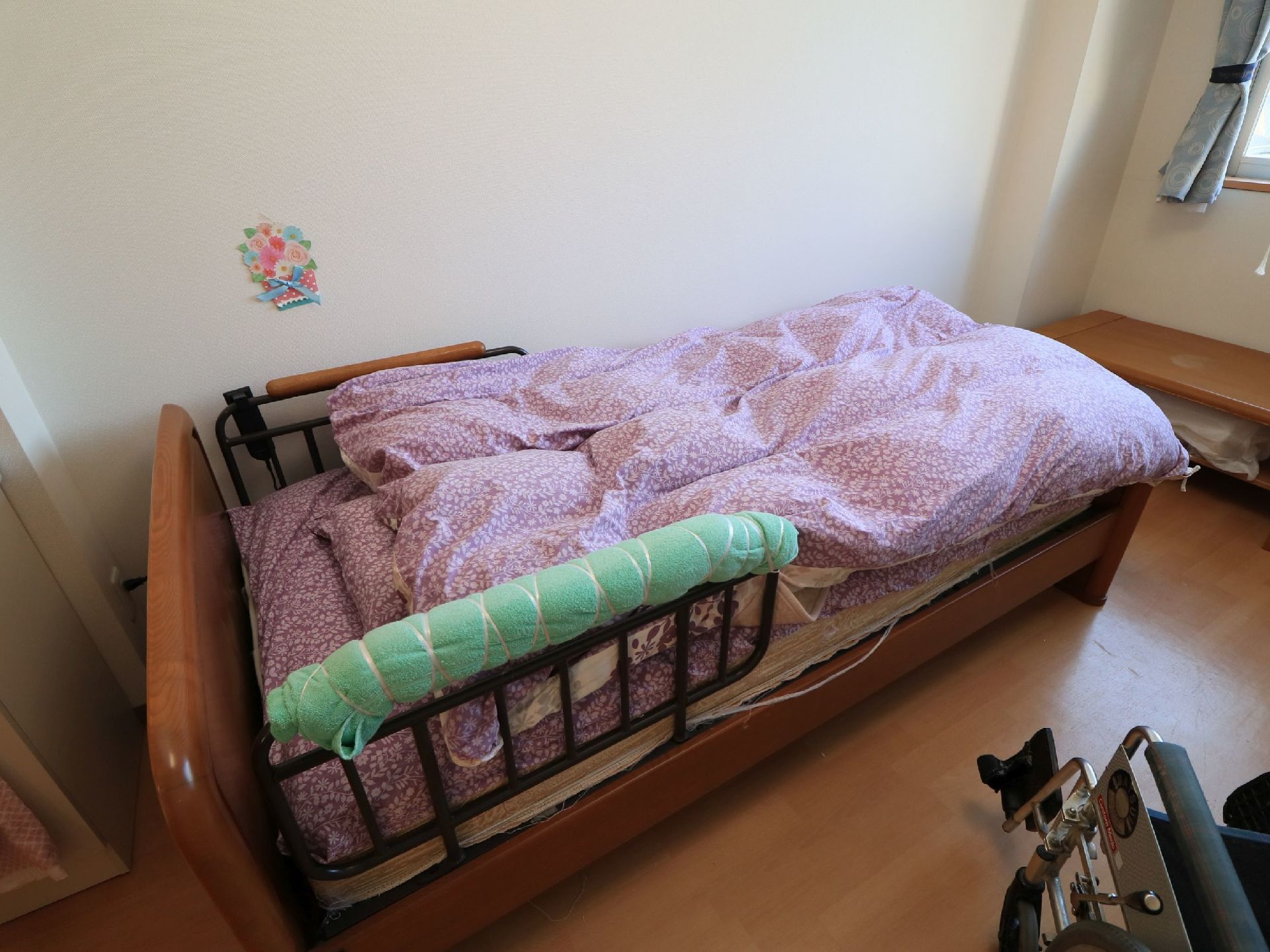 壁際にベッドが設置されている。ピンクの花柄の布団が置かれていて、手すり部分にグリーンのタオルが巻かれている。