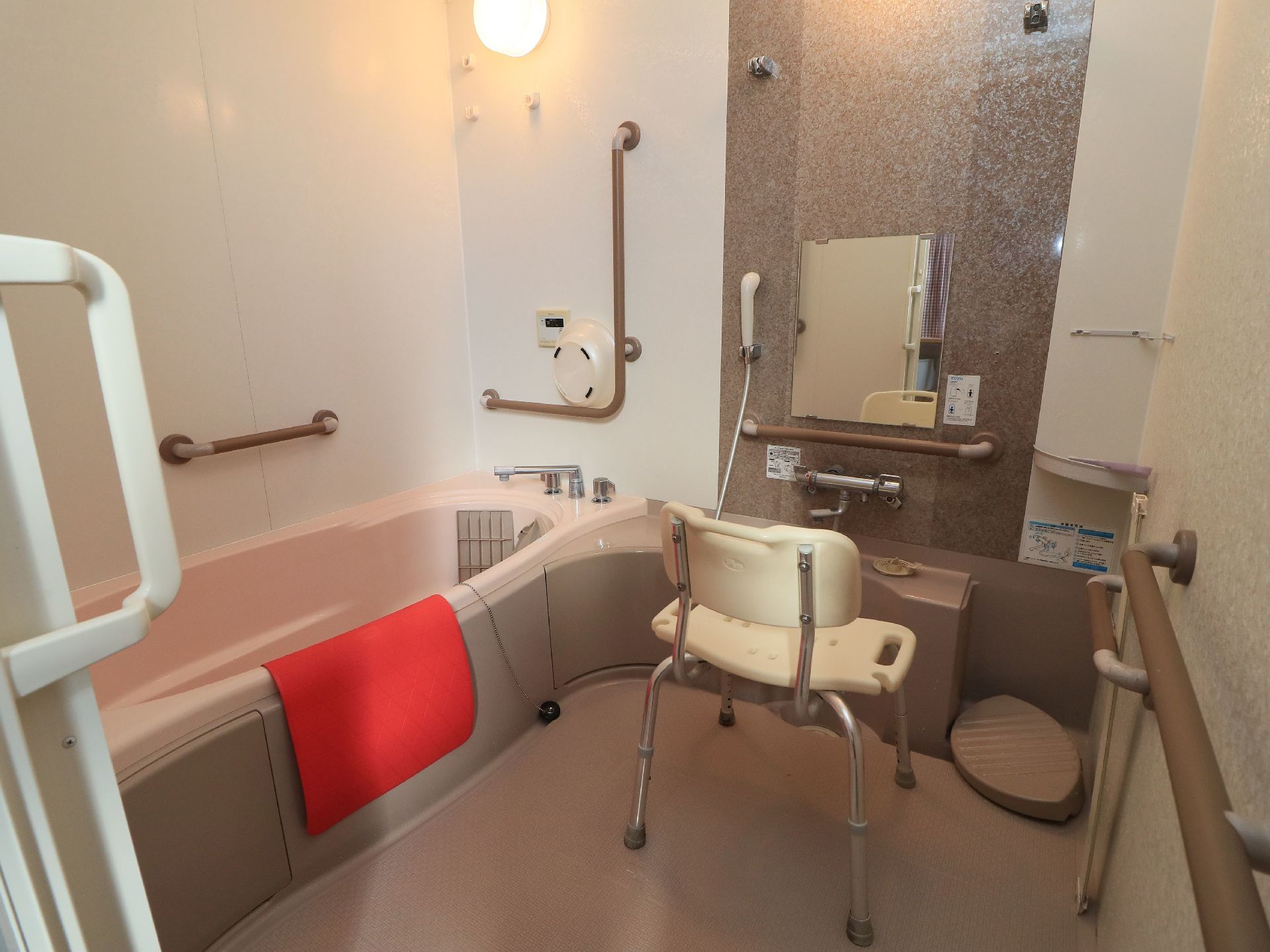 ブラウンを基調とした浴室で、白いシャワーチェアが置かれている。バスタブの縁には赤いマットがかけられている。