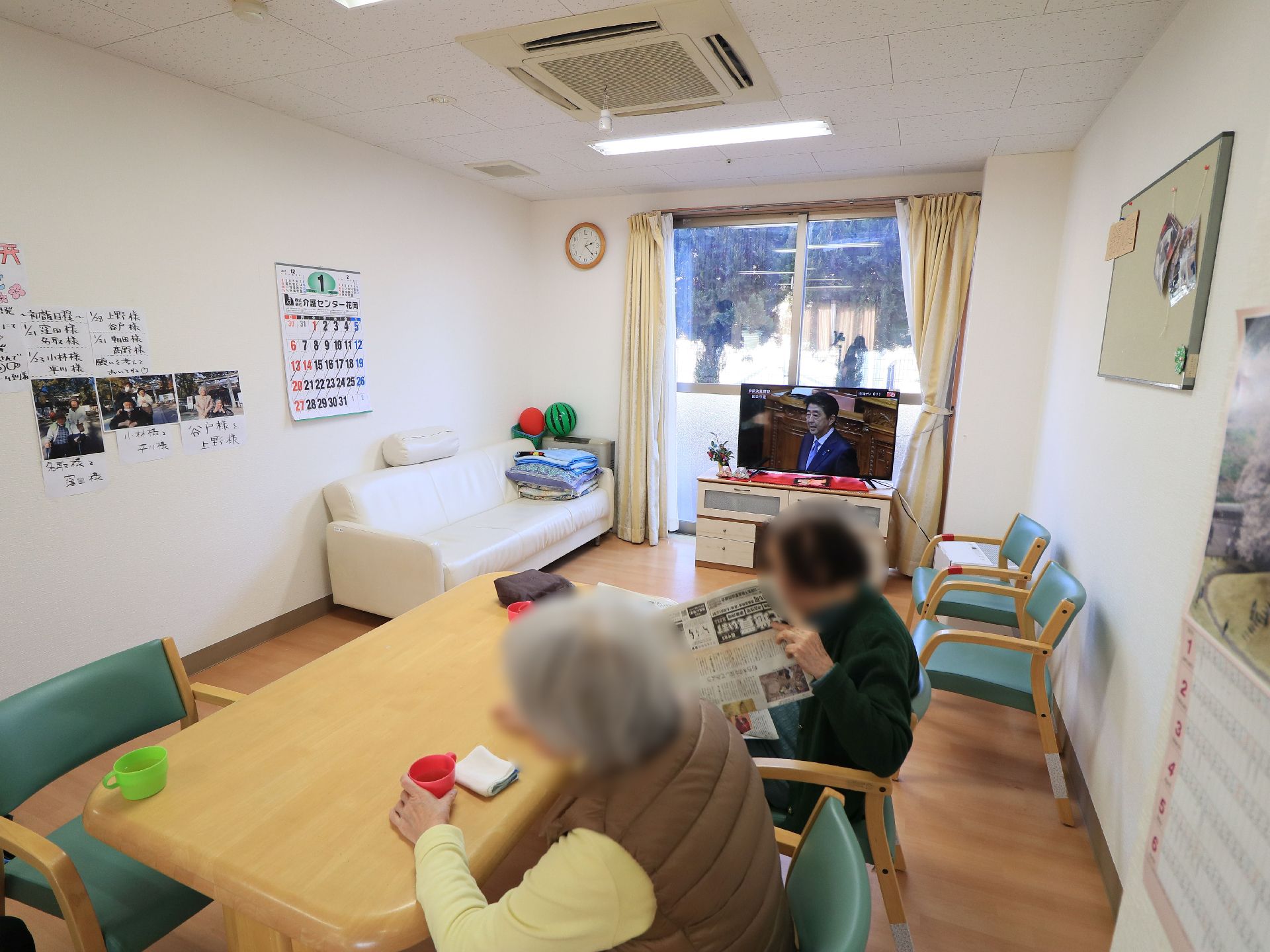 食堂で入居者様が２人並んで座っている。１人は新聞を読んでいて、もう１人は飲み物のコップを手にしている。