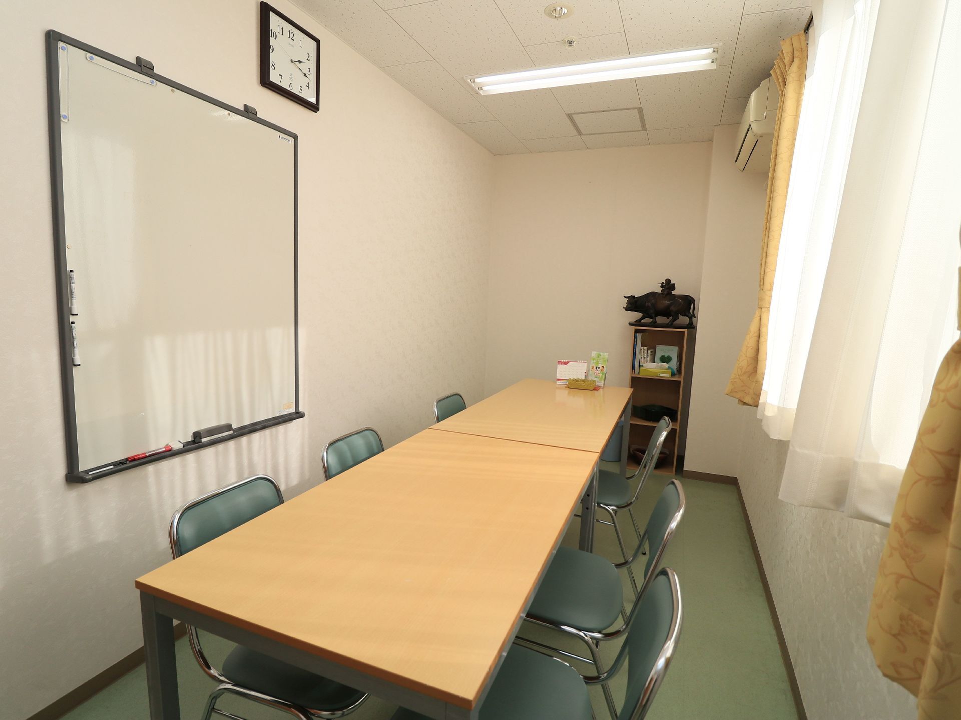 長方形のテーブルが２つくっつけて置かれ、グリーンの椅子が６脚配置されている。壁にホワイトボードがかけられている。