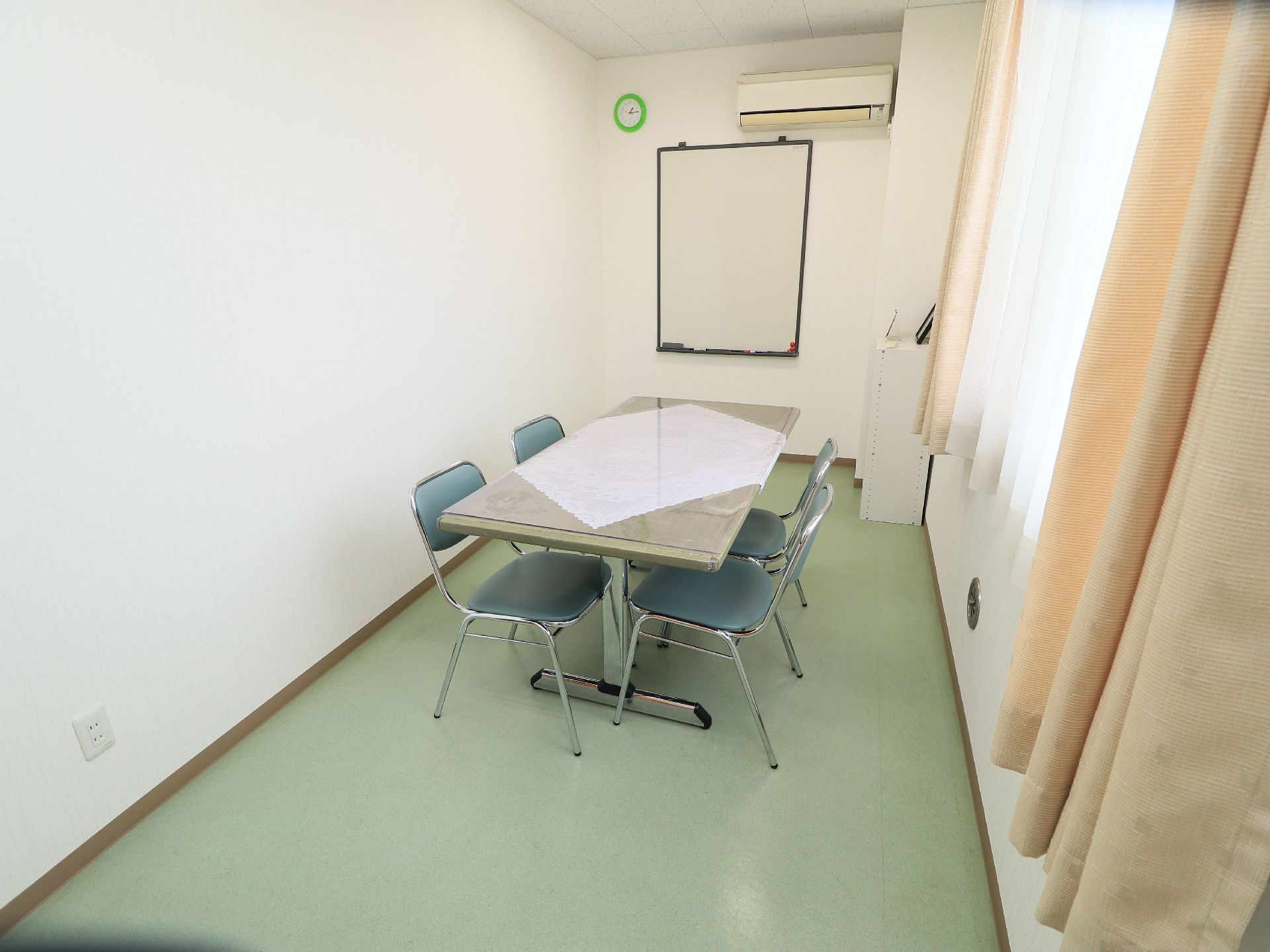 長方形のテーブルとグリーンの椅子が４脚置かれている。部屋の奥の壁にはホワイトボードと時計、エアコンが設置されている。