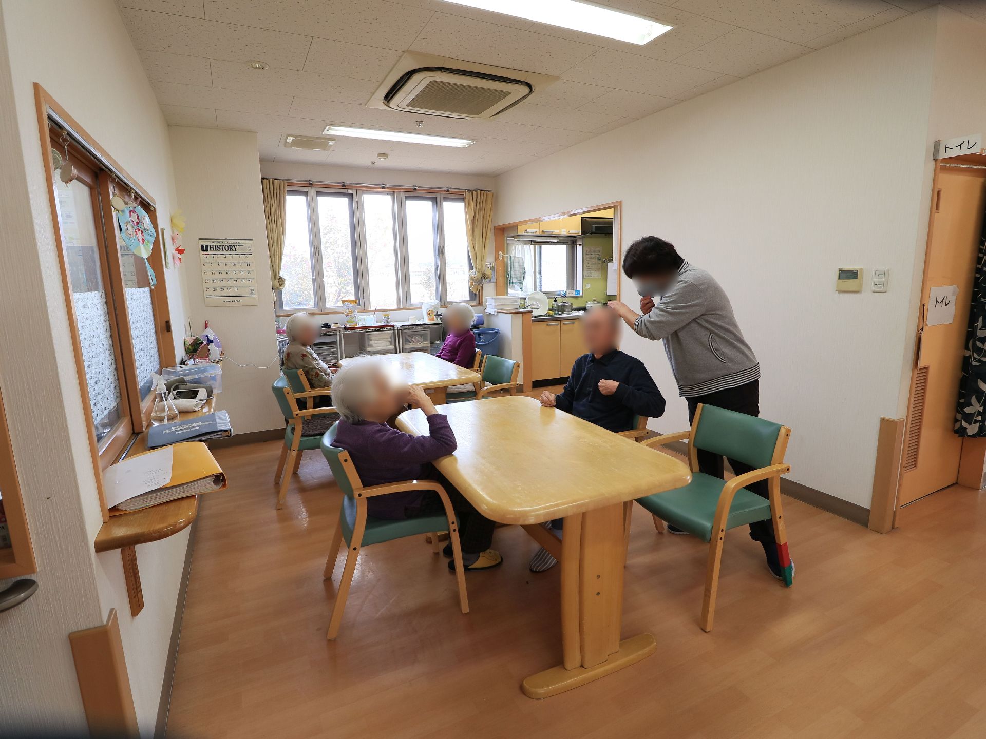 食堂で４人の入居者様が椅子に座っている。１人の入居者様の横にスタッフが立ち、手を差し出して会話を交わしている。