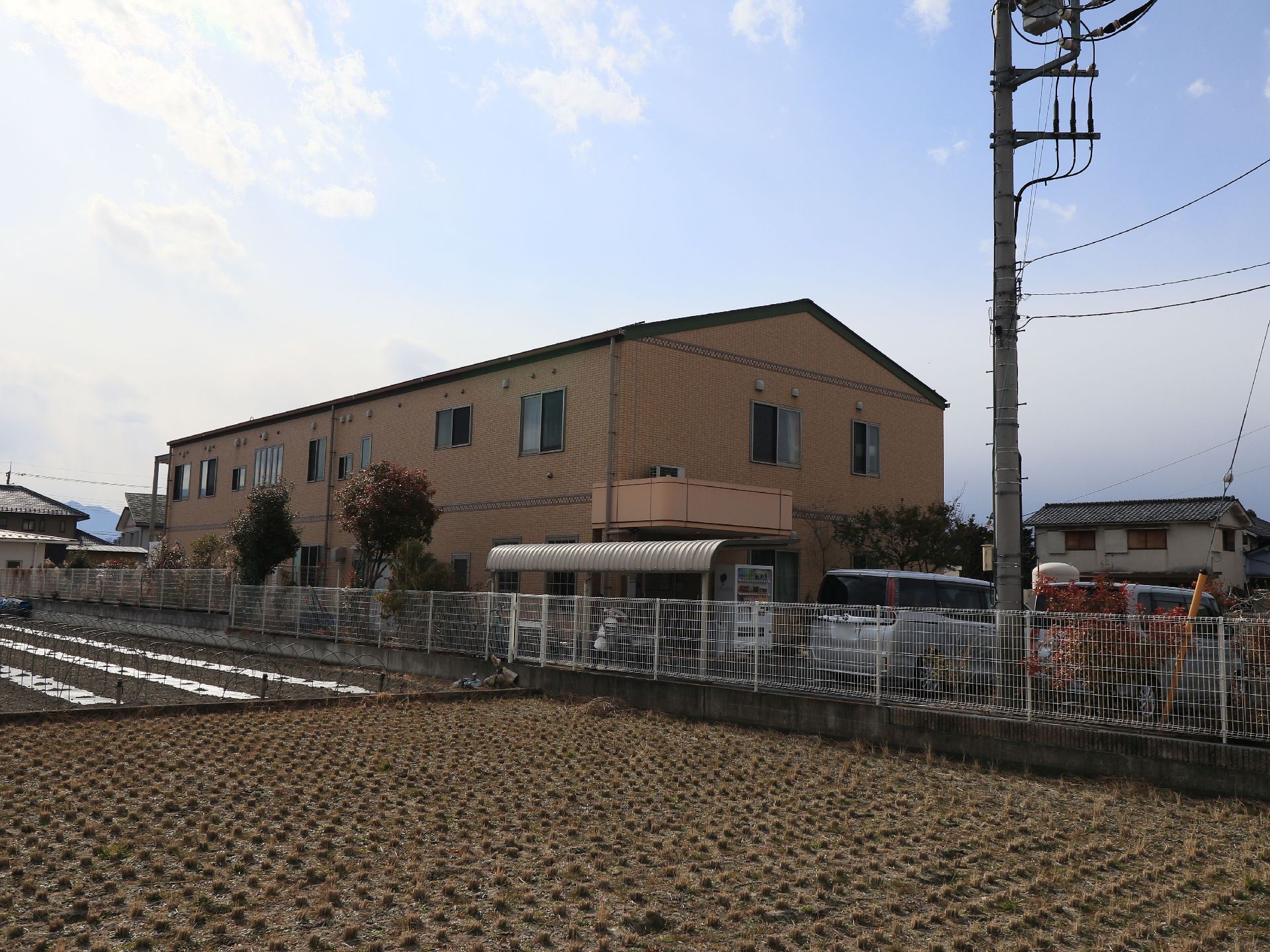 建物のすぐ横には広い田んぼと畑があり、周囲には住宅があるのが見える。敷地の境界には白いフェンスが設置されている。