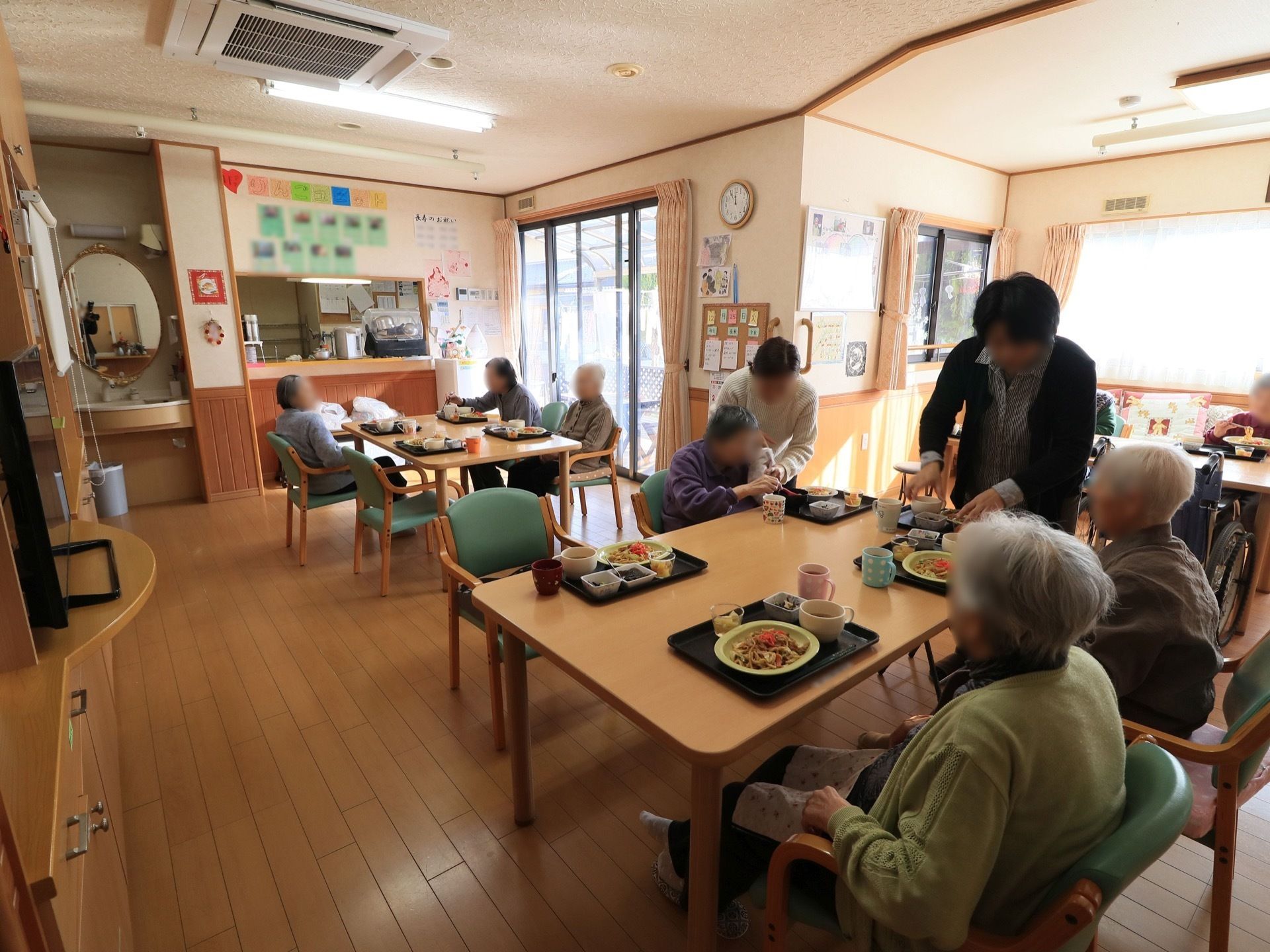 ４人掛けのテーブルとひじ掛けのついた椅子が並べられている食堂で、入居者様がお食事を召し上がっている。カウンターキッチンがある。
