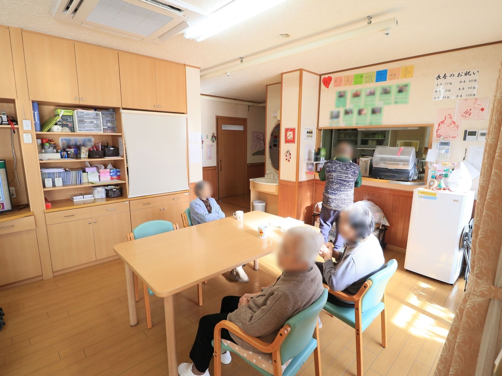 食堂にテーブルと椅子が置かれ、入居者様がお茶を一服されている。カウンターキッチンがある。壁一面が棚になっており、日常使う物が収納されている。