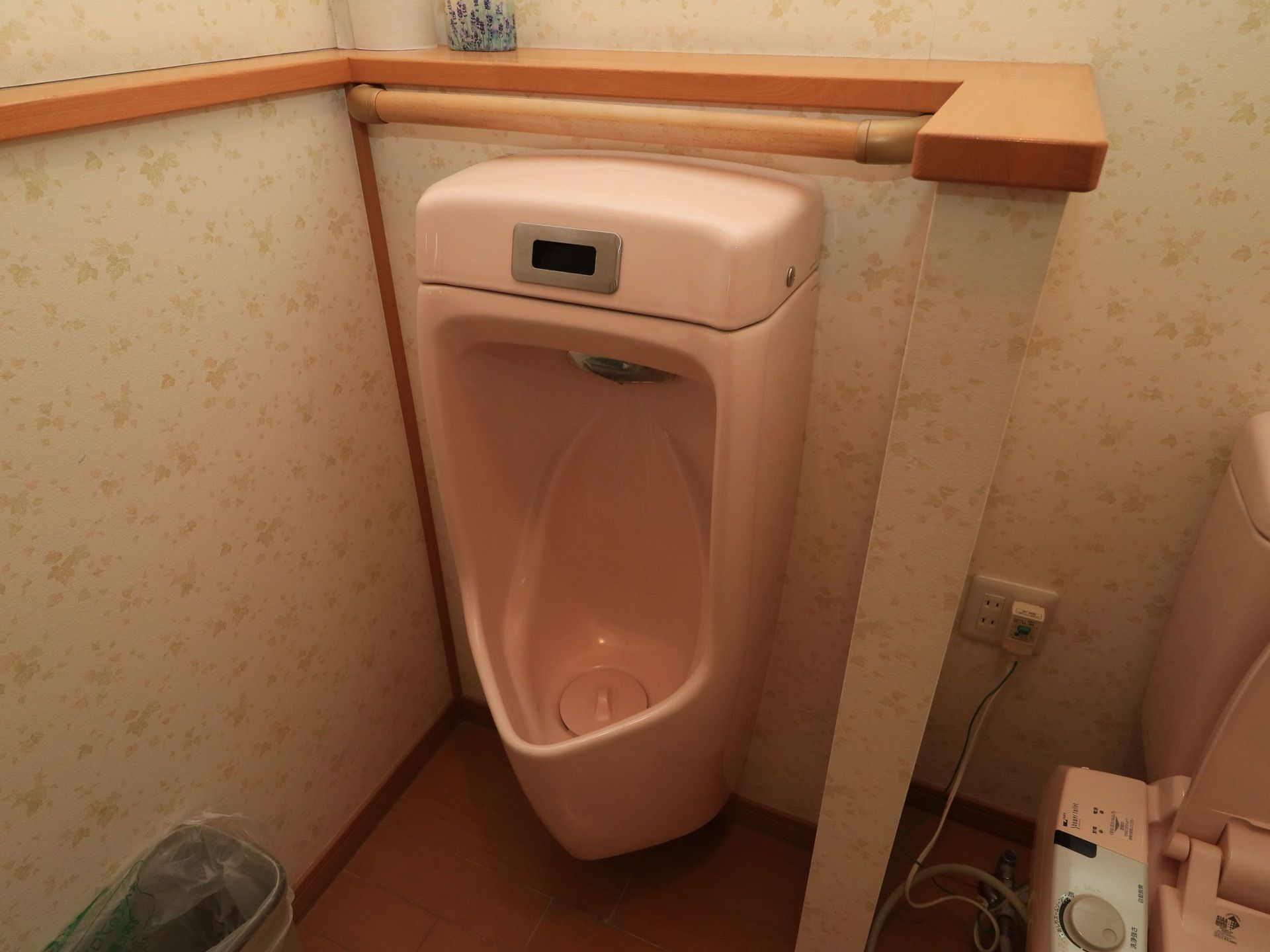 花柄の壁紙の壁で、ピンク色の男性用の便器が置かれている。周りに仕切り壁があり、手すりが設置されている。