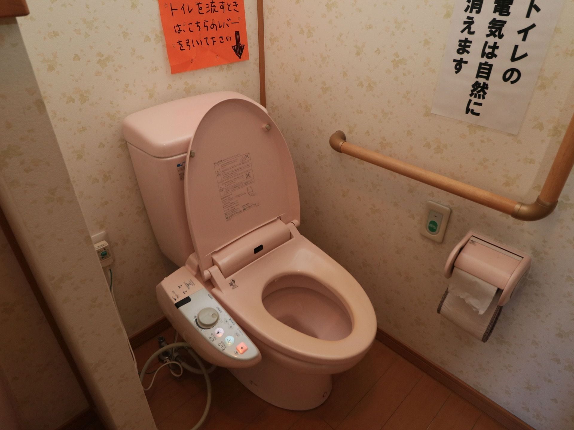 ピンク色の洋式便器が設置されている。花柄の壁には縦横方面に手すりが取りつけられており、注意書きの張り紙が貼られている。