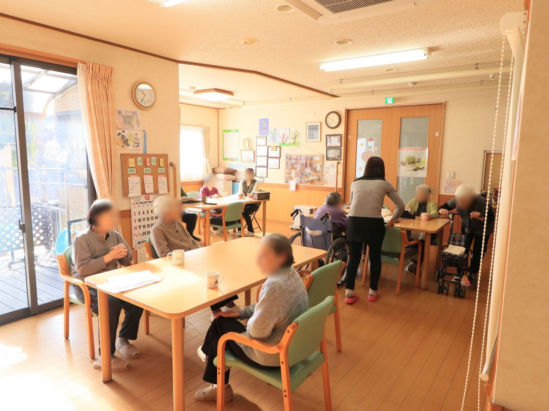食堂には大人数で座れるテーブルと椅子が置かれている。バルコニーに続く大きな窓がある。壁にカレンダーやメモが貼られたコルクボードが掛っている。