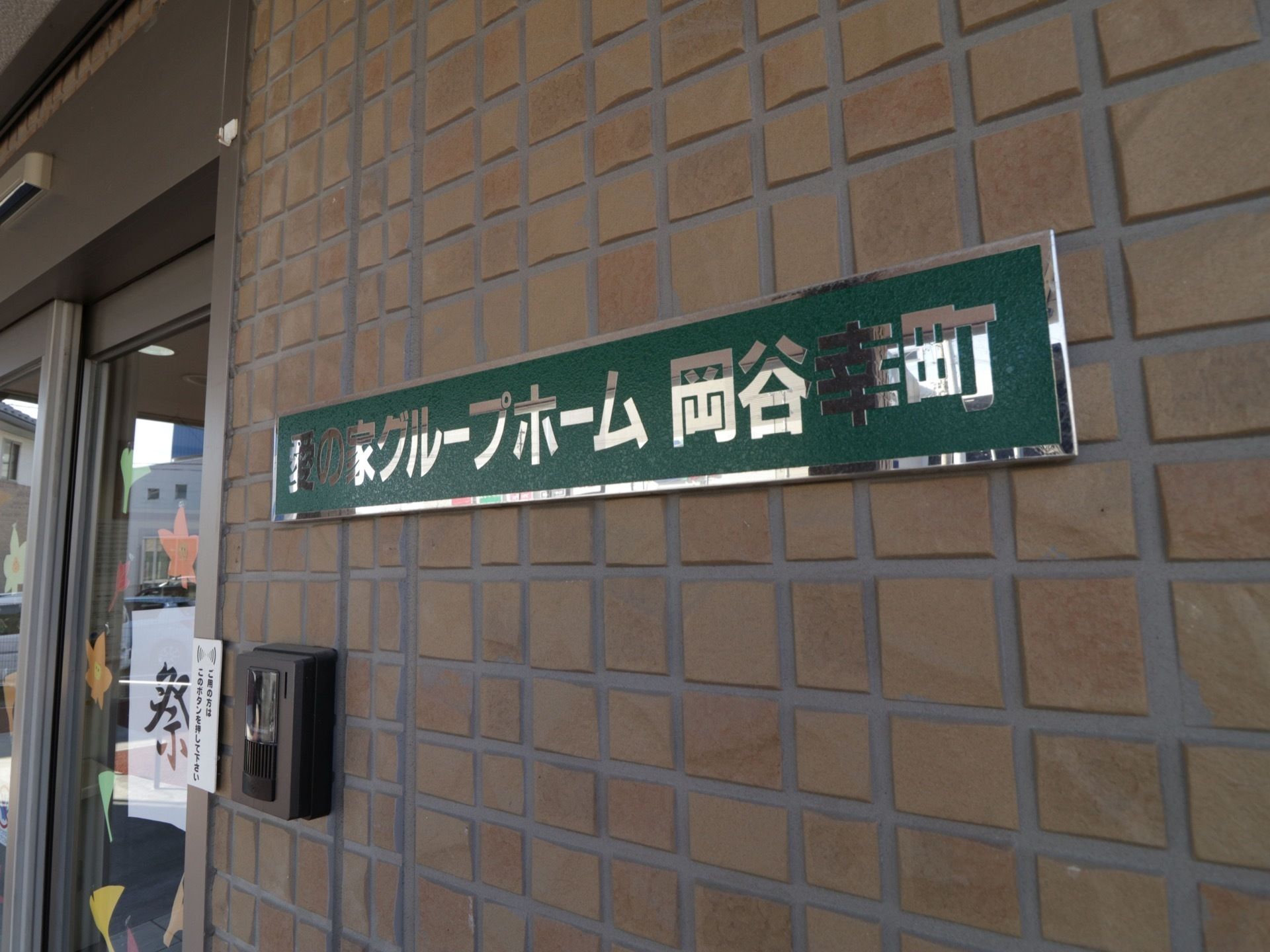 玄関横の外壁に看板が取りつけられている。深い緑色ベースにシルバーの縁取りで、施設名が書かれている。建物に馴染んだ見やすい看板である。