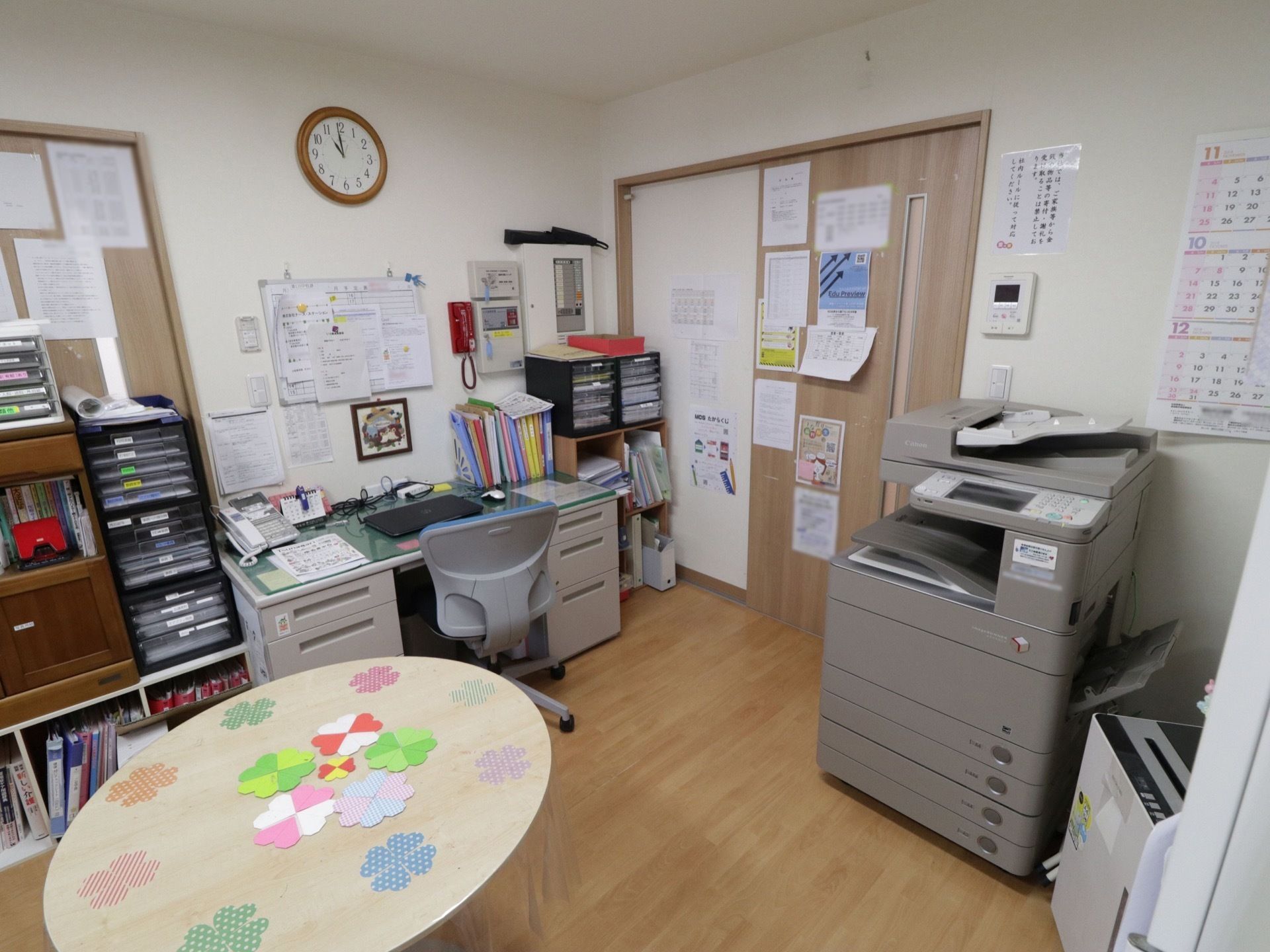 パソコンが用意されたデスクがあり、その周囲には書類棚が多数置かれている。ドアの右側にコピー機が設置されている。