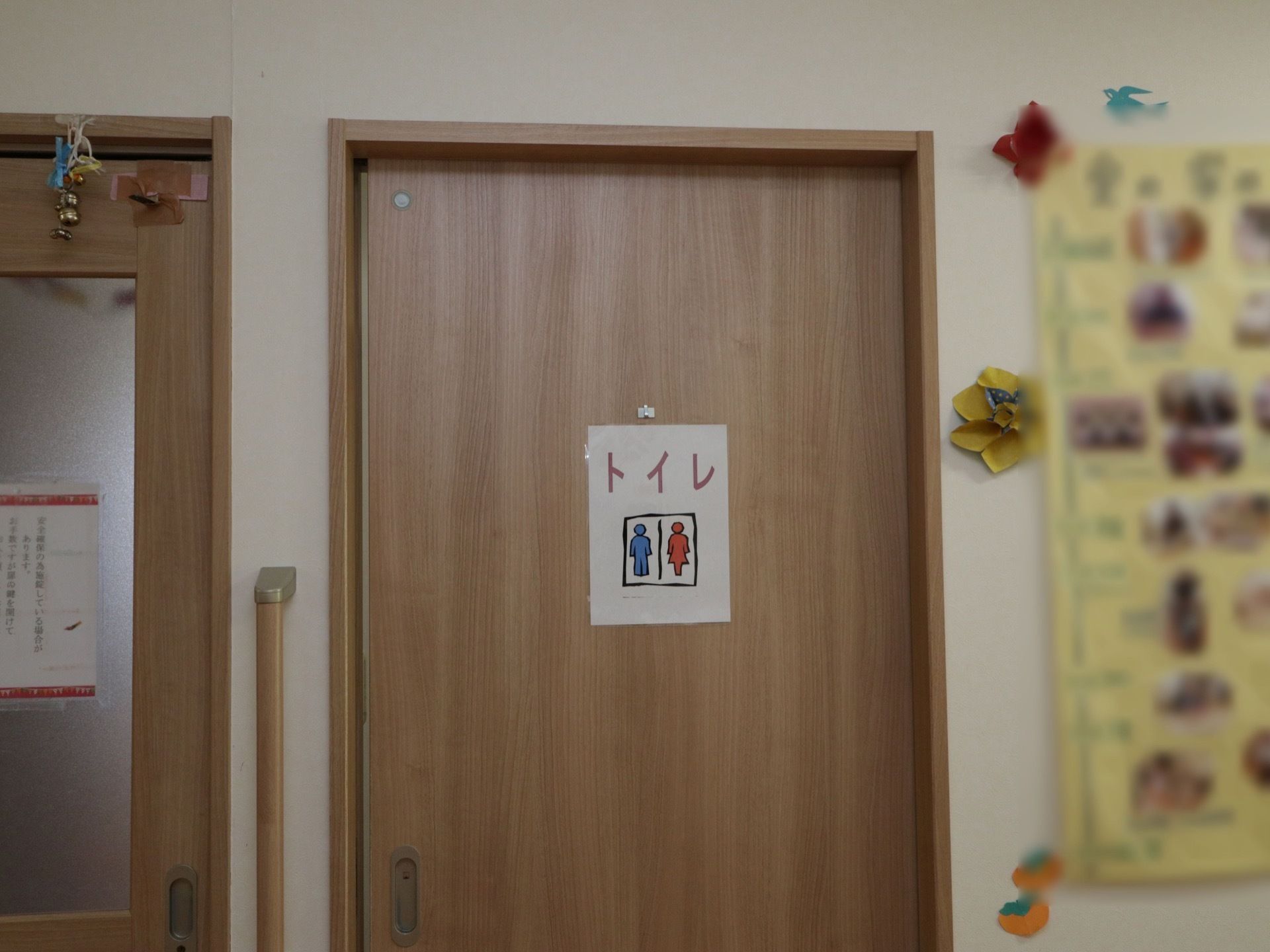スライド式のドアの共用トイレで、ドアの左側に縦長の手すりが設置されている。右側の壁には、模造紙に写真を貼り掲示している。