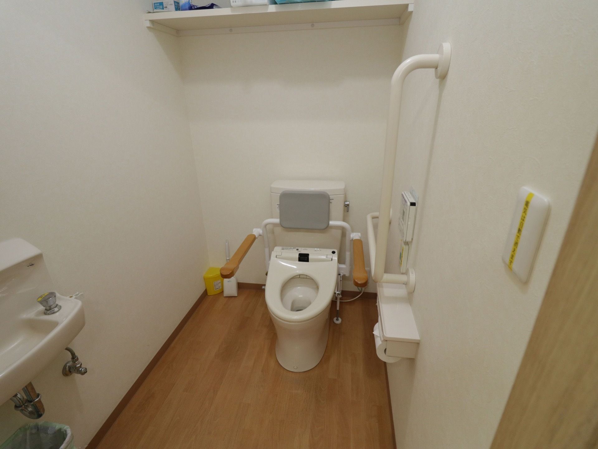 フローリングの床の個室トイレで、可動式のアームレストが設置された洋式便座がある。壁にはナースコールが取りつけられている。