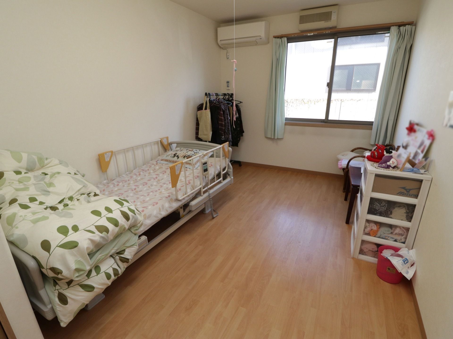 明るい色合いのフローリングと大きな窓がある居室。ベッドは介護用で、小さい棚と椅子、ハンガーラックが置かれている。