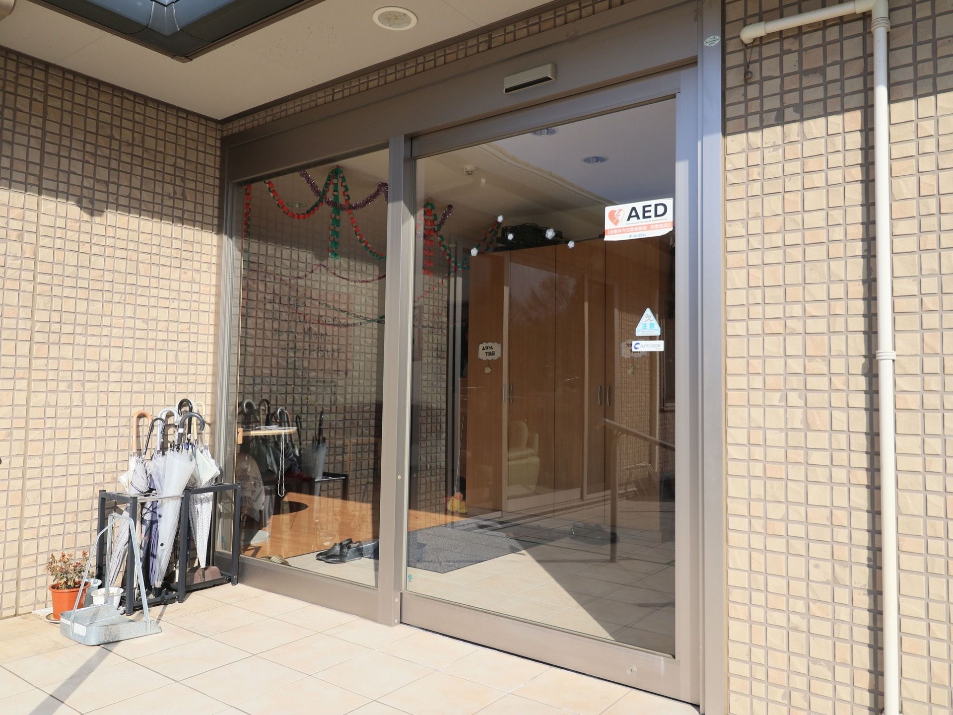 ガラス張りの自動ドアになっている。ドアにはAEDのシールが貼られており、玄関の前には傘立てが置かれている。