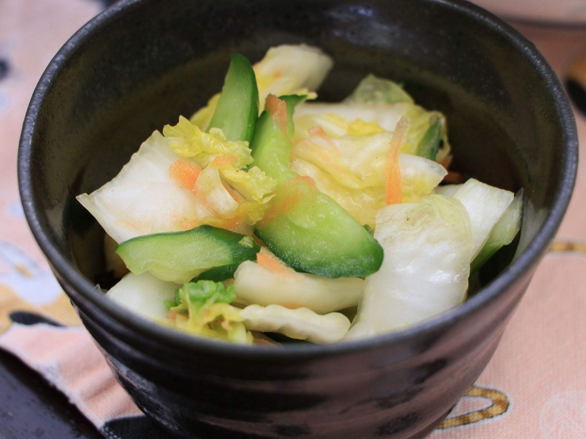 黒い陶器の皿の中に白菜、きゅうりの漬物が入っている。少々、にんじんも添えられており、彩りが鮮やかな一品になっている。