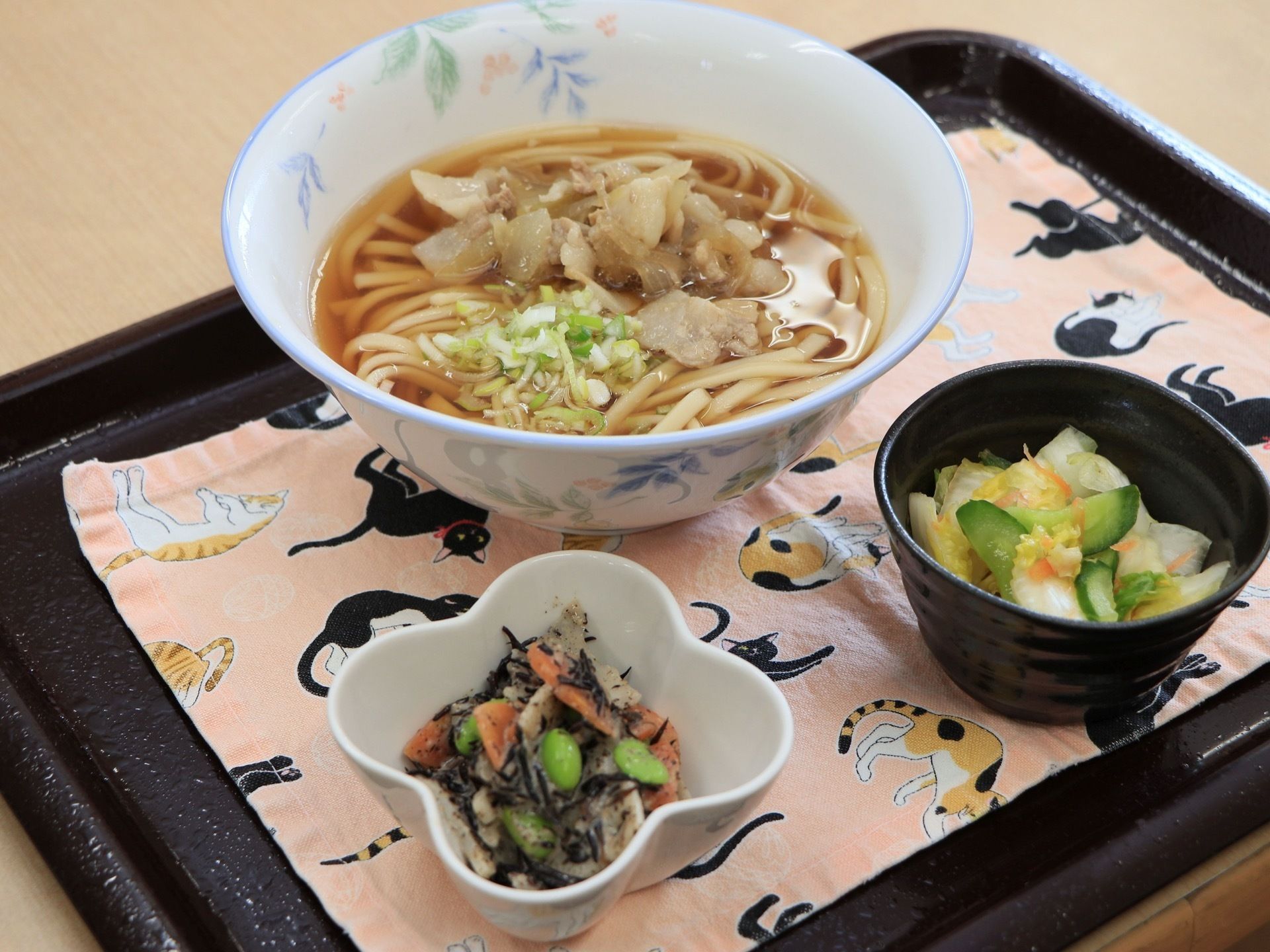 猫柄の可愛らしいランチョマットが敷かれている。うどんをメインにお漬物が添えられており、バランスの良い食事になっている。