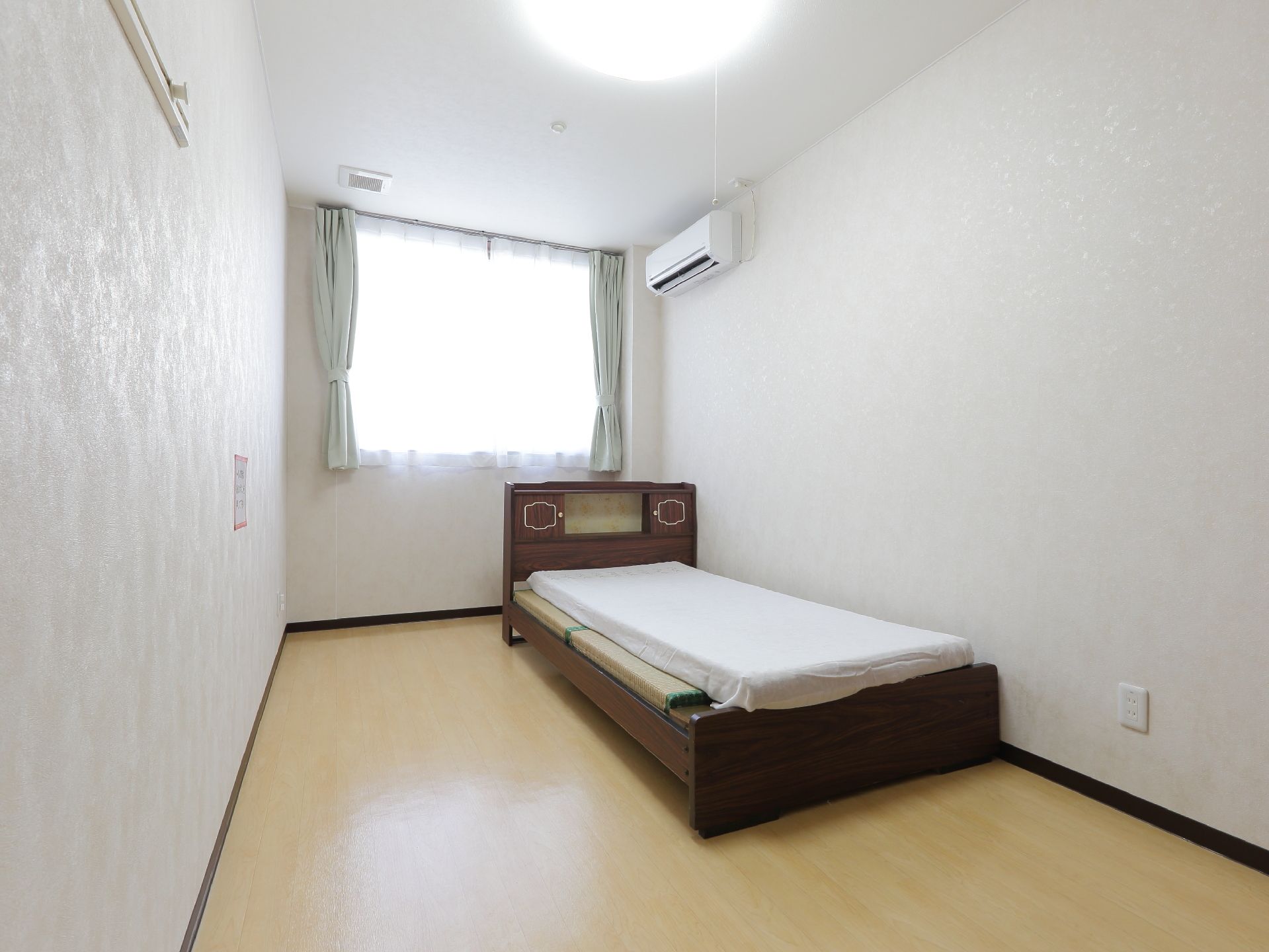 居室は縦長で、ベッド、ライトグリーンのカーテンとレースカーテン、エアコンが設置されている。壁には衣紋掛もある。