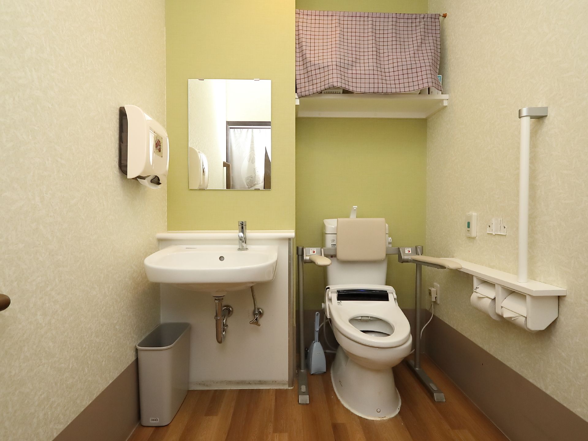 明るいグリーンの壁色が印象的なトイレである。壁には緊急呼び出しボタン、縦型の手すりが設けられている。