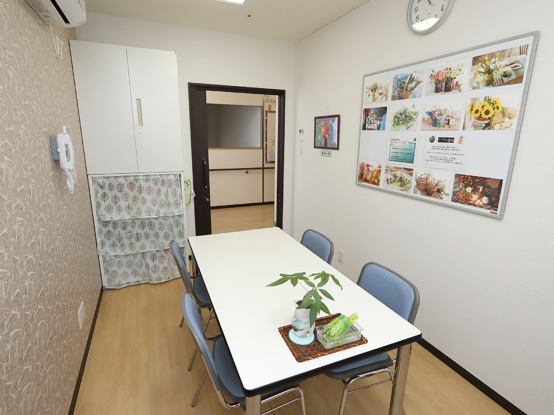 白色のテーブルに、ブルーの背もたれ椅子が４脚置かれている。観葉植物が飾られている。エアコンも備えつけられている。