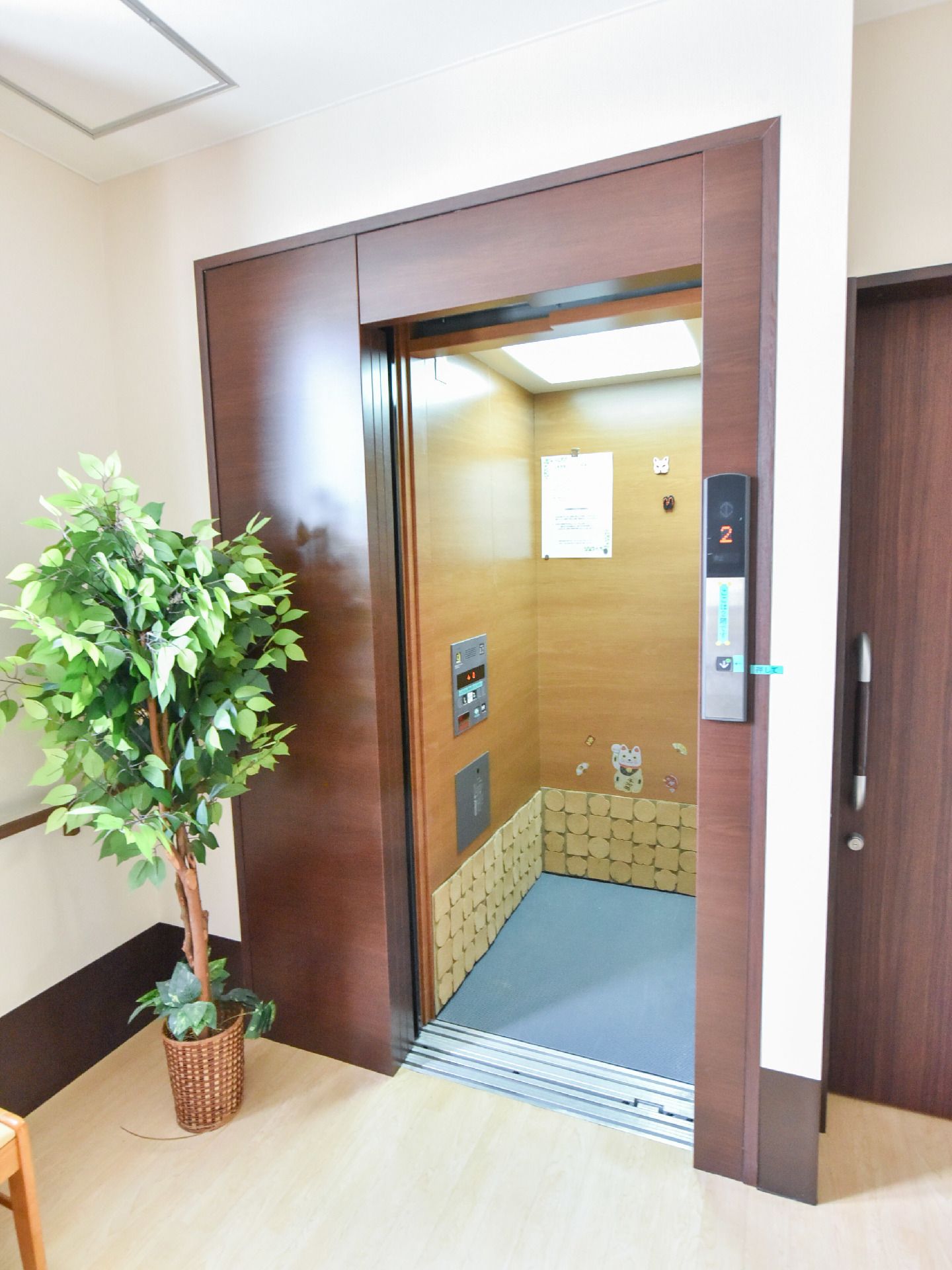 エレベーターの中の壁には招き猫などの柄が付いている他、張り紙がされている。木目の壁のエレベーターである。