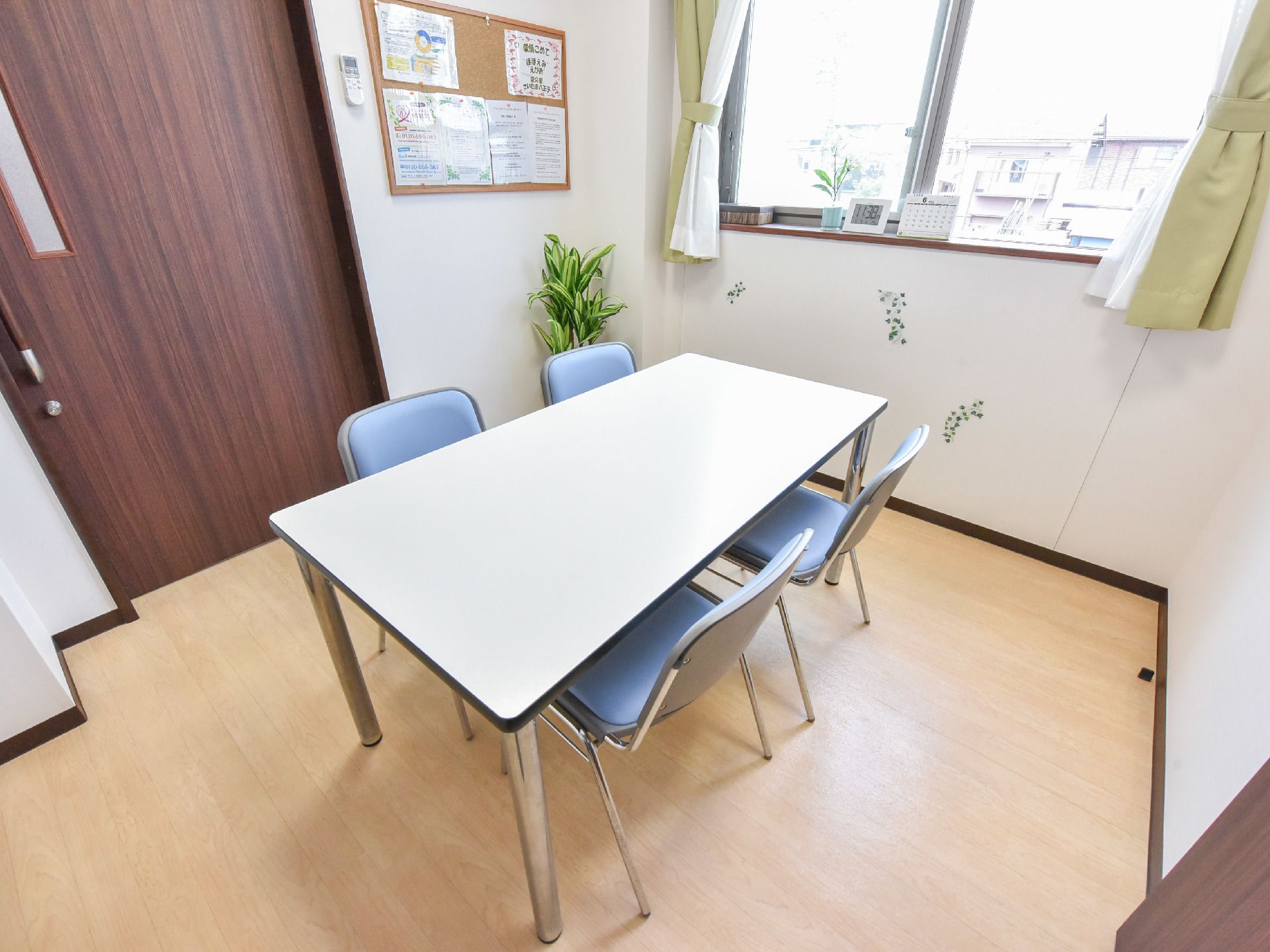 小さな個室に4人がけのテーブルと机がある。窓枠には時計とカレンダーが置かれている。部屋の隅に観葉植物がある。
