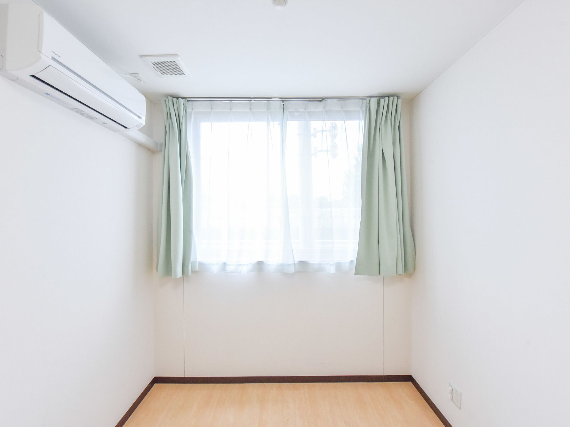 薄いグリーンのカーテンが白い壁とナチュラル色のフローリングに映える室内である。エアコンが設置されている。