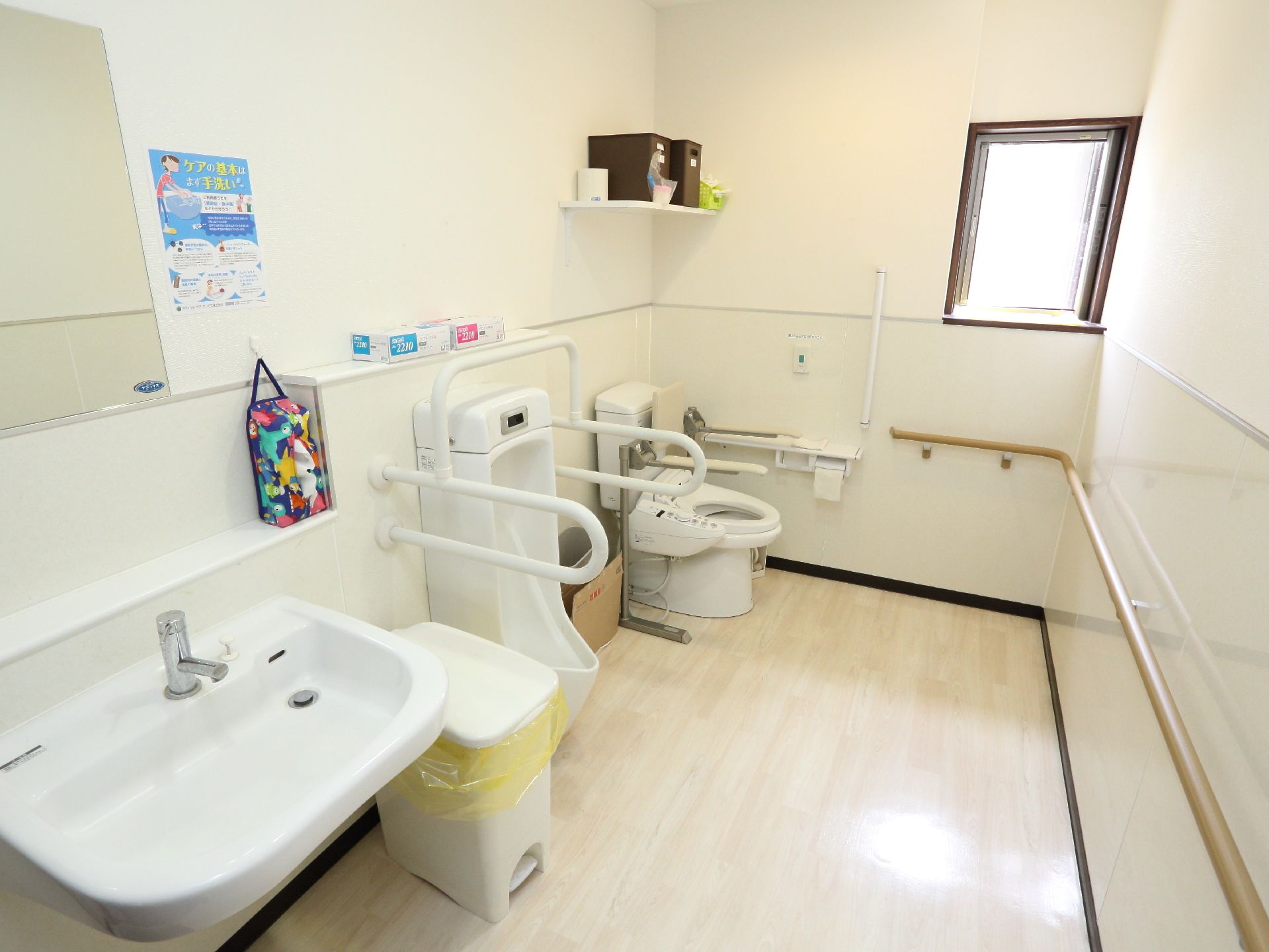 自動洗浄器付きトイレ、男性用の便器、手洗い場が直線に並んでいる。壁には手すりが付いている。それぞれの便器にも手すりが付けられている。