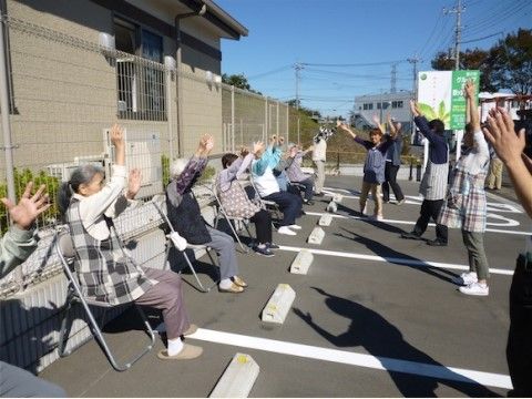 駐車場でパイプ椅子に座りながら上半身の運動に勤しむシニア