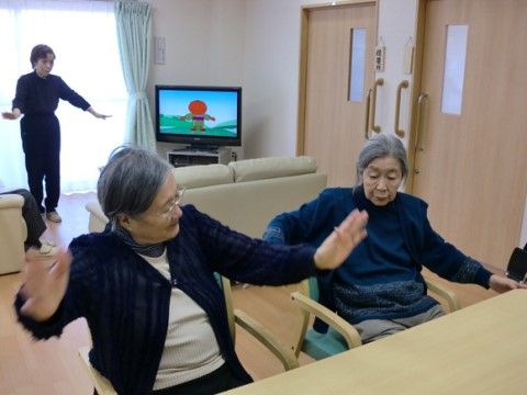 椅子に座りながらＴＶの音声に合わせて運動を楽しむシニア女性