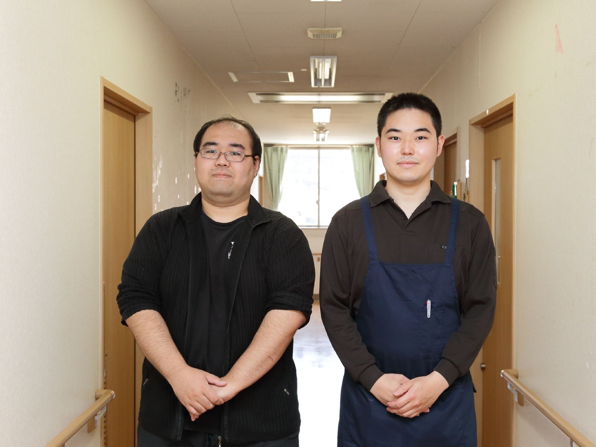 突き当りに窓のある白い壁の廊下に並んで立つ二人の男性