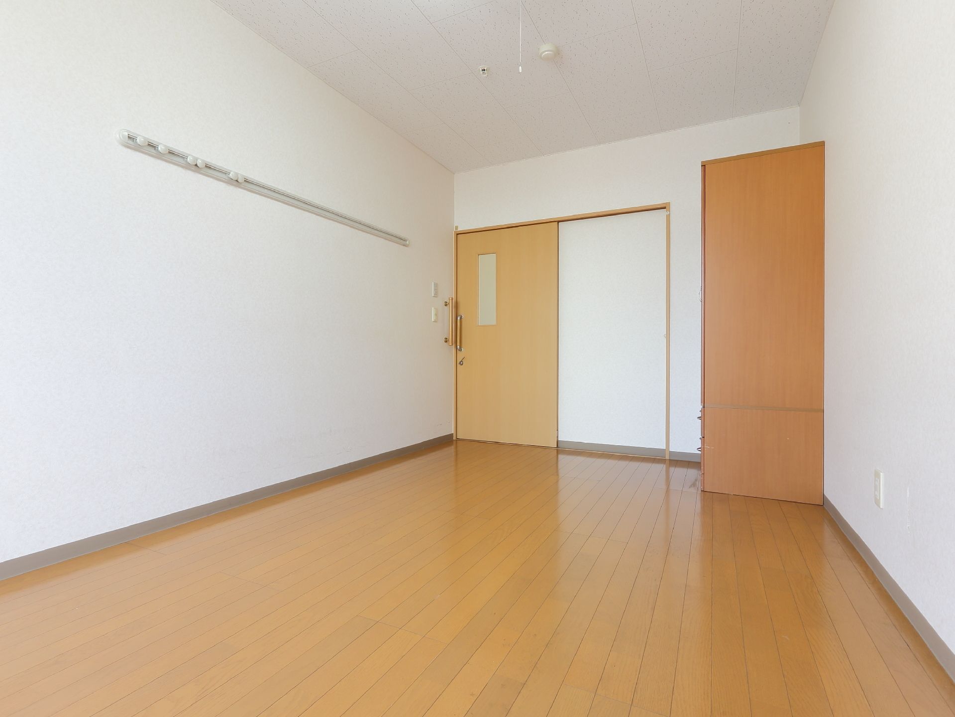 白と茶色のナチュラル色で落ち着いた居室。入り口脇にはクローゼットが備わっている。天井には火災報知機が設置されている。