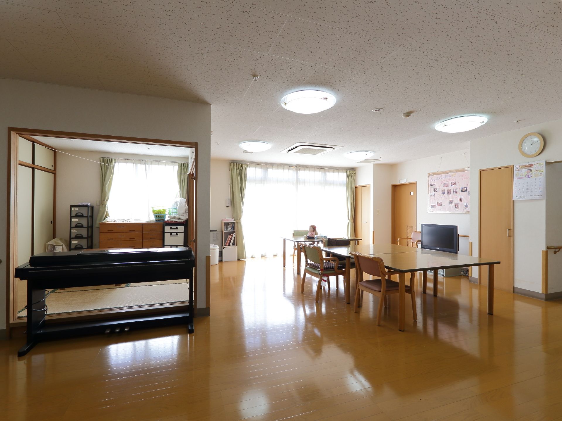 大きな窓のある明るい共有リビングの様子。広々とした空間にテーブルと椅子、ワイド型のテレビ、電子ピアノなどが置かれている。