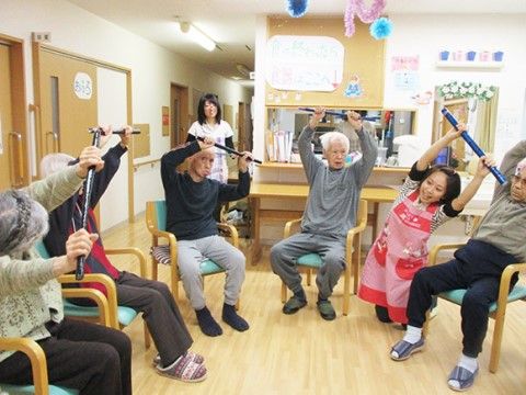 椅子で輪になって、体操を行っている。手作りの棒を使ってスタッフの見本を見ながら、腕や上半身を伸ばしている。