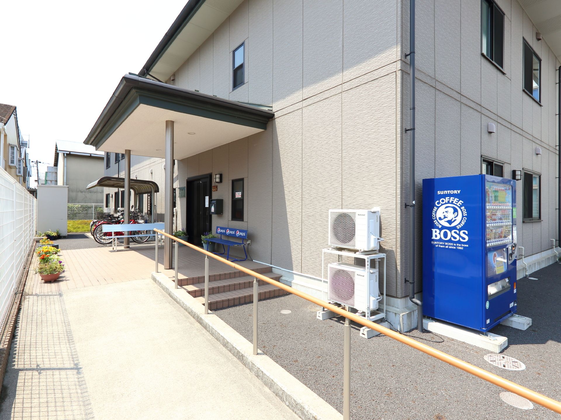 建物に通じる手すり付の通路があります。玄関の入口ドアの所に2つのベンチが置いてある。外壁角側に自動販売機がある。