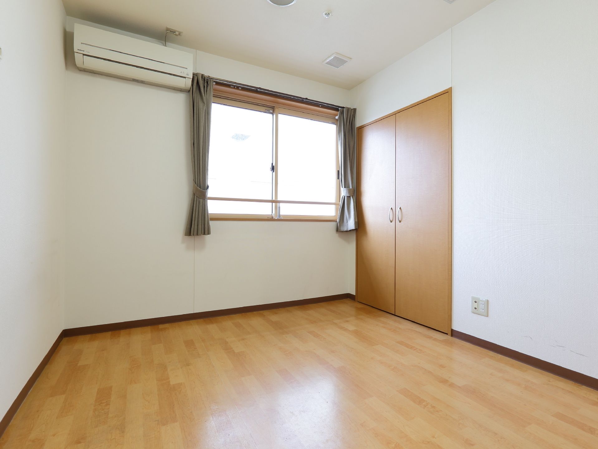 白色の壁と天井、木目調の床に窓が設置。エアコンの設備も整っている。窓傍の広い開き戸があり、その横下にコンセントがある。