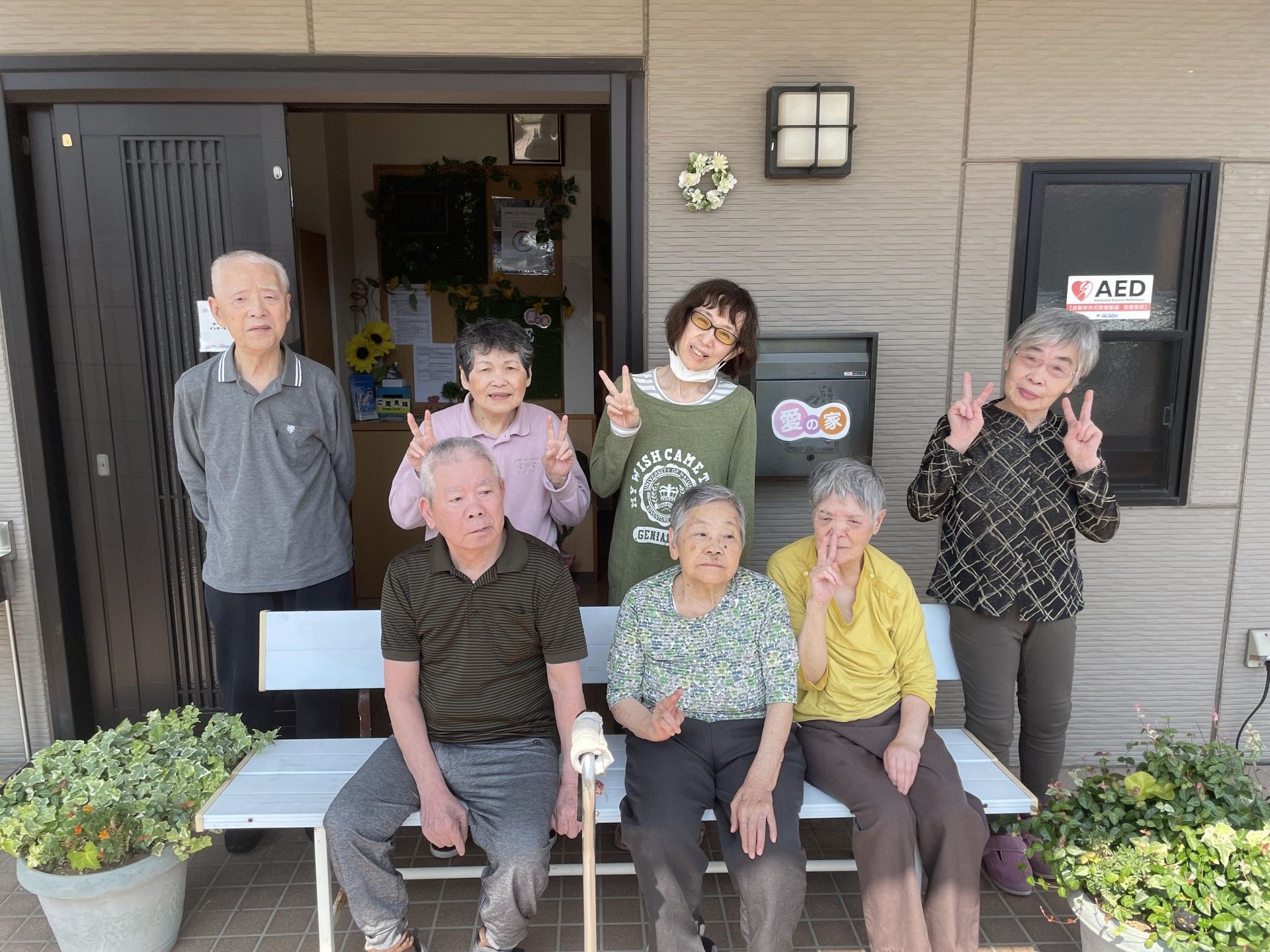 玄関の外で記念撮影をする５人のシニアと３人のヘルパー