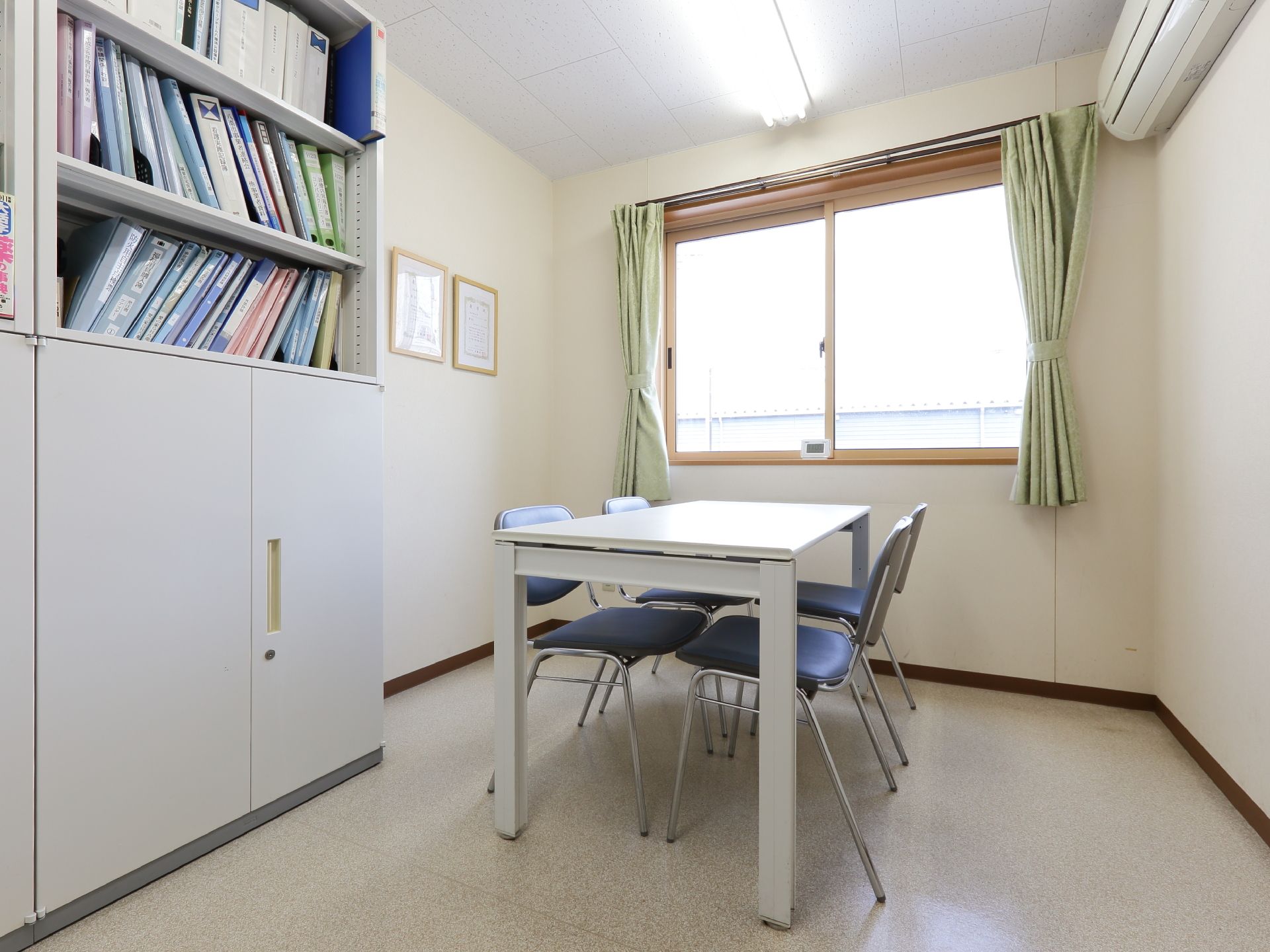白で統一された内装の一室。正面には、木枠の窓があり、テーブルと椅子が4つ置いてある。手前には、ファイルなどの収納棚が置いてある。